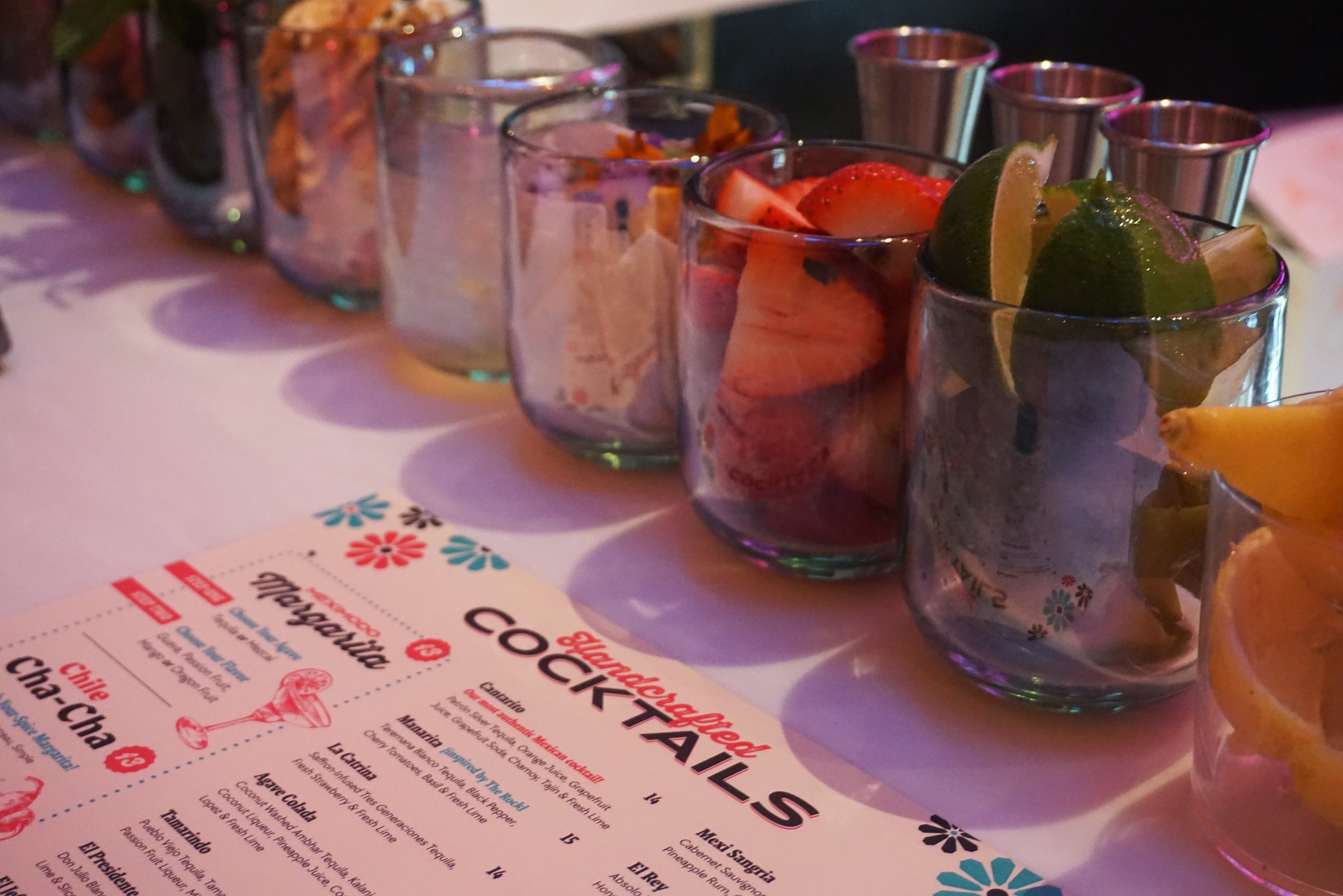Row of glass cups filled with sliced fruits like strawberries, lemons, and limes on a table, with a colorful menu visible in the foreground.