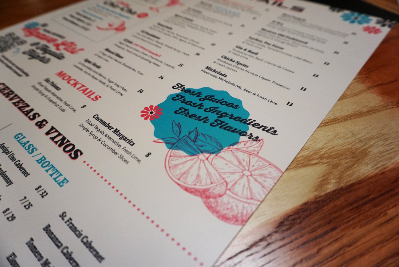 Close-up of a colorful menu featuring fresh juices, fruit ingredients, cocktails, and beverages, with a decorative illustration of citrus slices and herbs on a wooden table.
