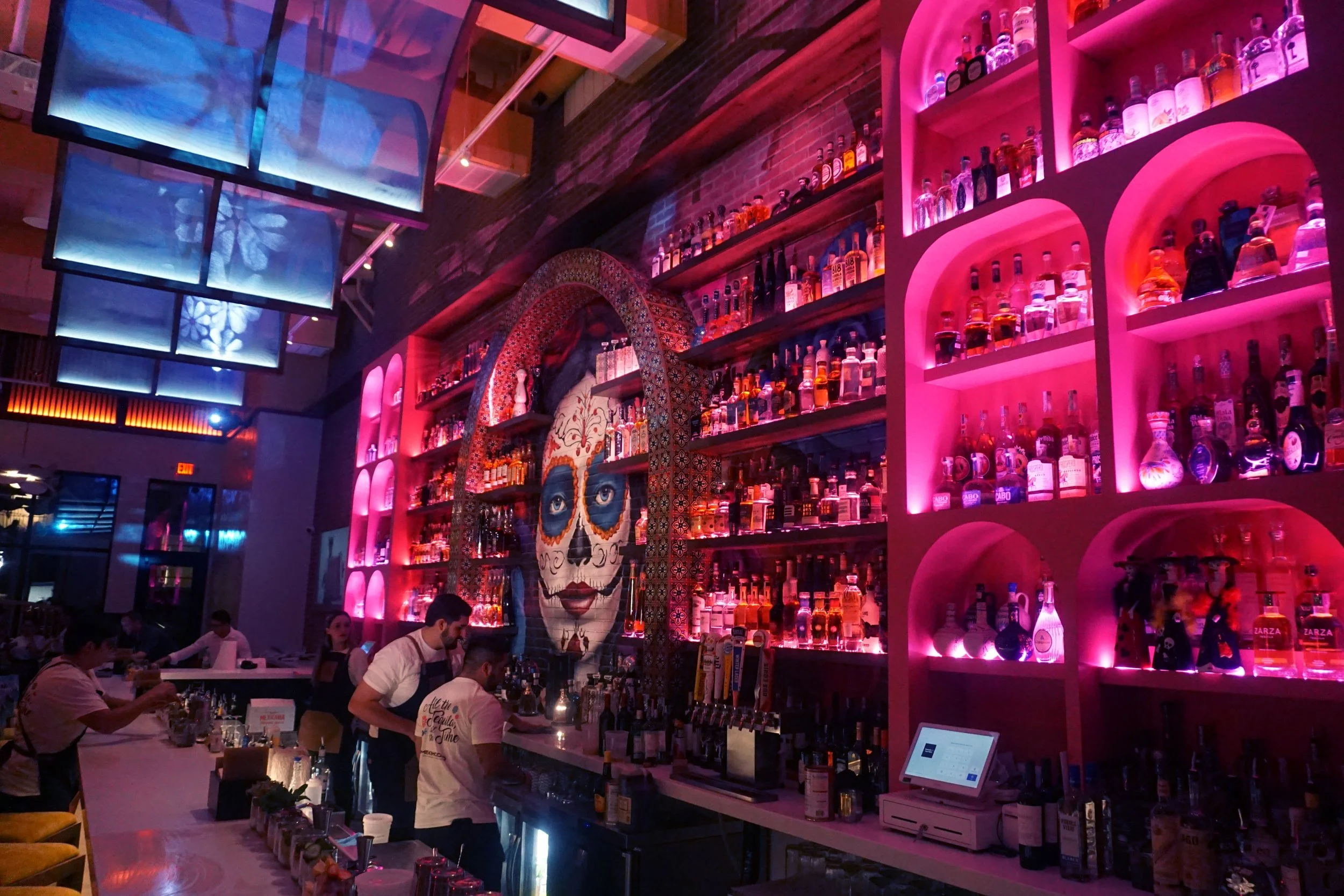 A lively bar with pink lighting, shelves filled with various bottles of alcohol, and a large decorative skull mural in the background. Bar staff are preparing drinks, and customers are seated at the bar.