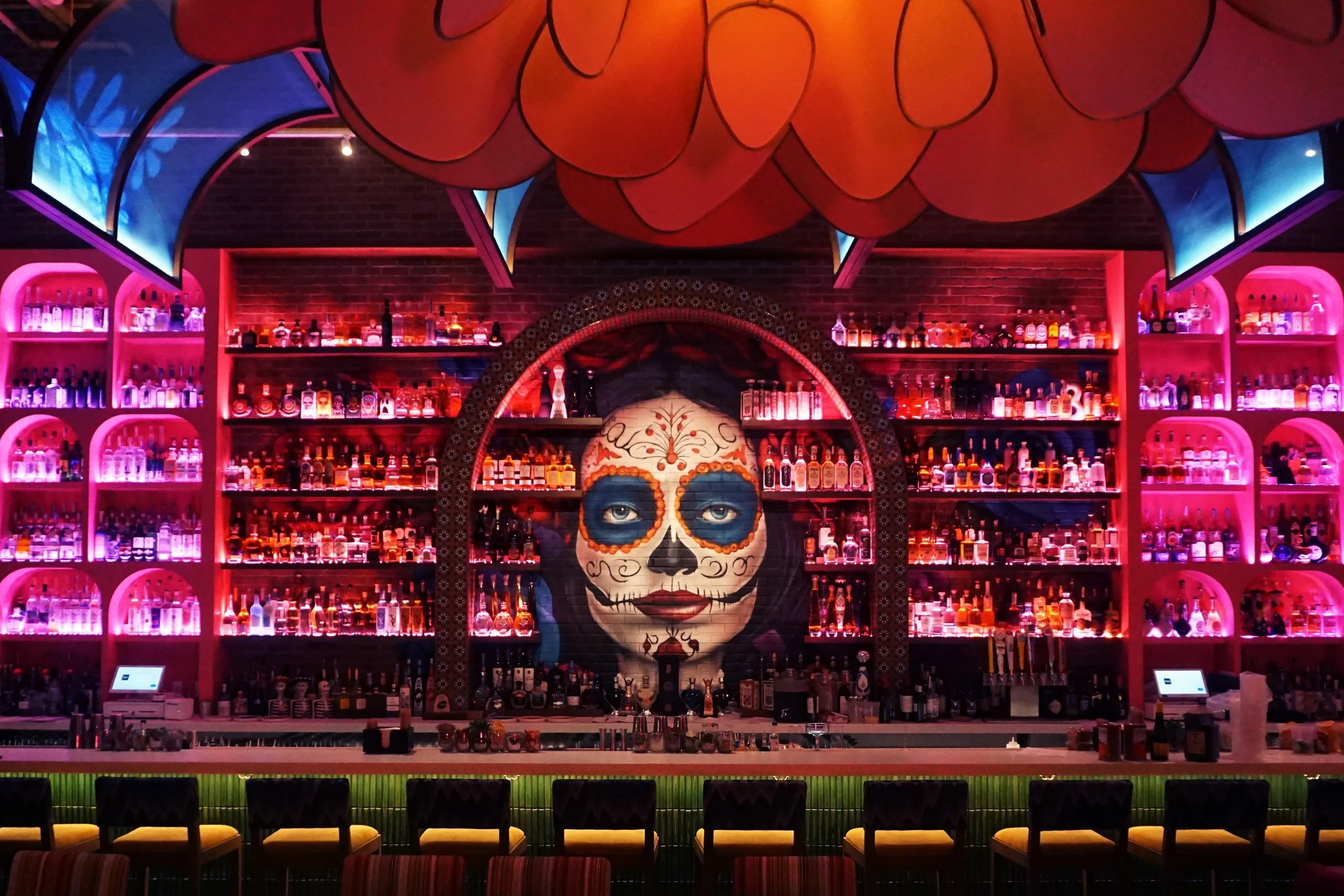 Colorful bar with a mural of a woman painted in a day of the dead style, surrounded by illuminated shelves of liquor bottles, and decorated with vibrant, neon-like lights.