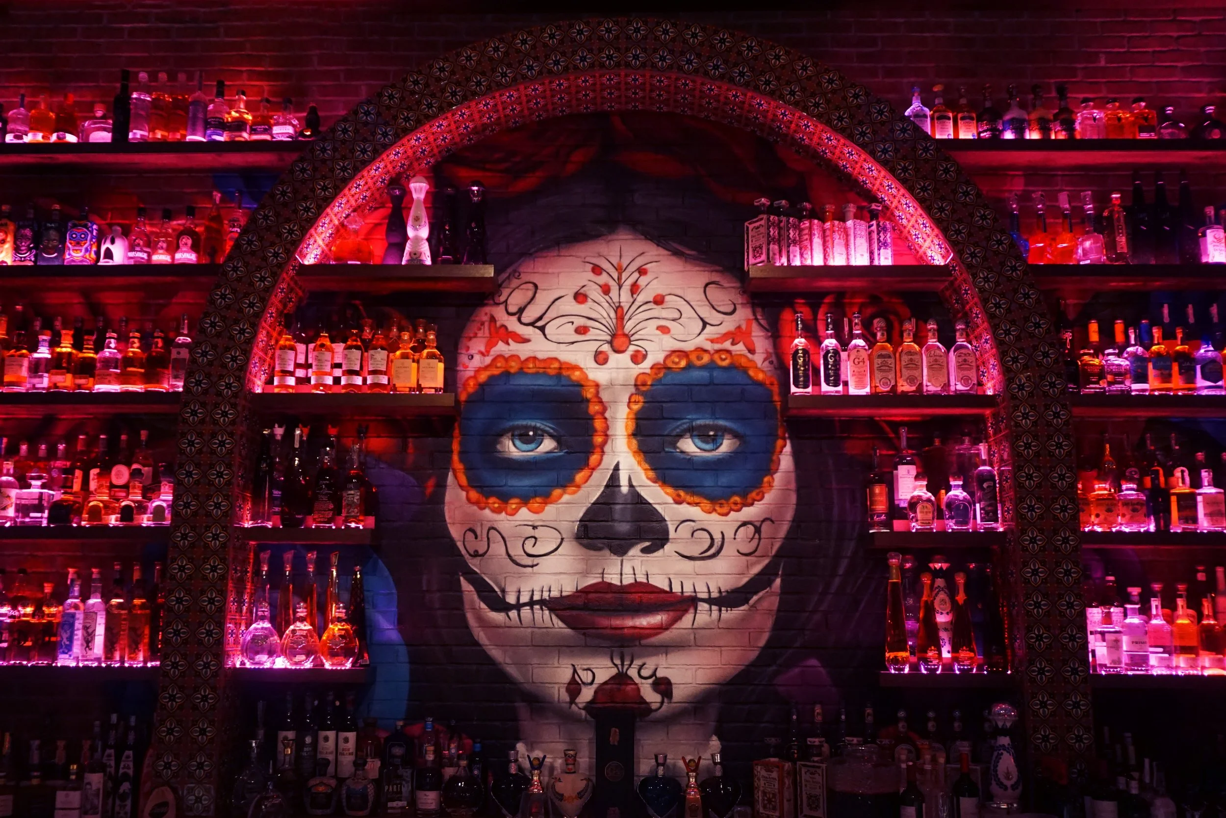 Colorful graffiti of a woman's face with Sugar Skull makeup, surrounded by illuminated liquor bottles on shelves.