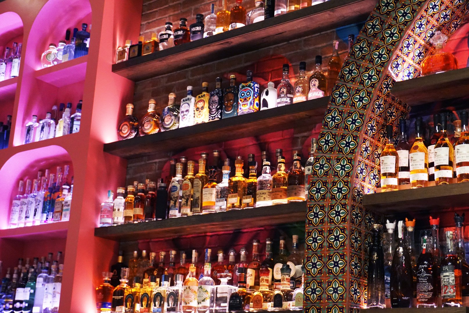 An indoor bar with shelves stocked with various bottles of alcohol, some with colorful labels and decorative caps, set against a brick wall with patterned tile details.