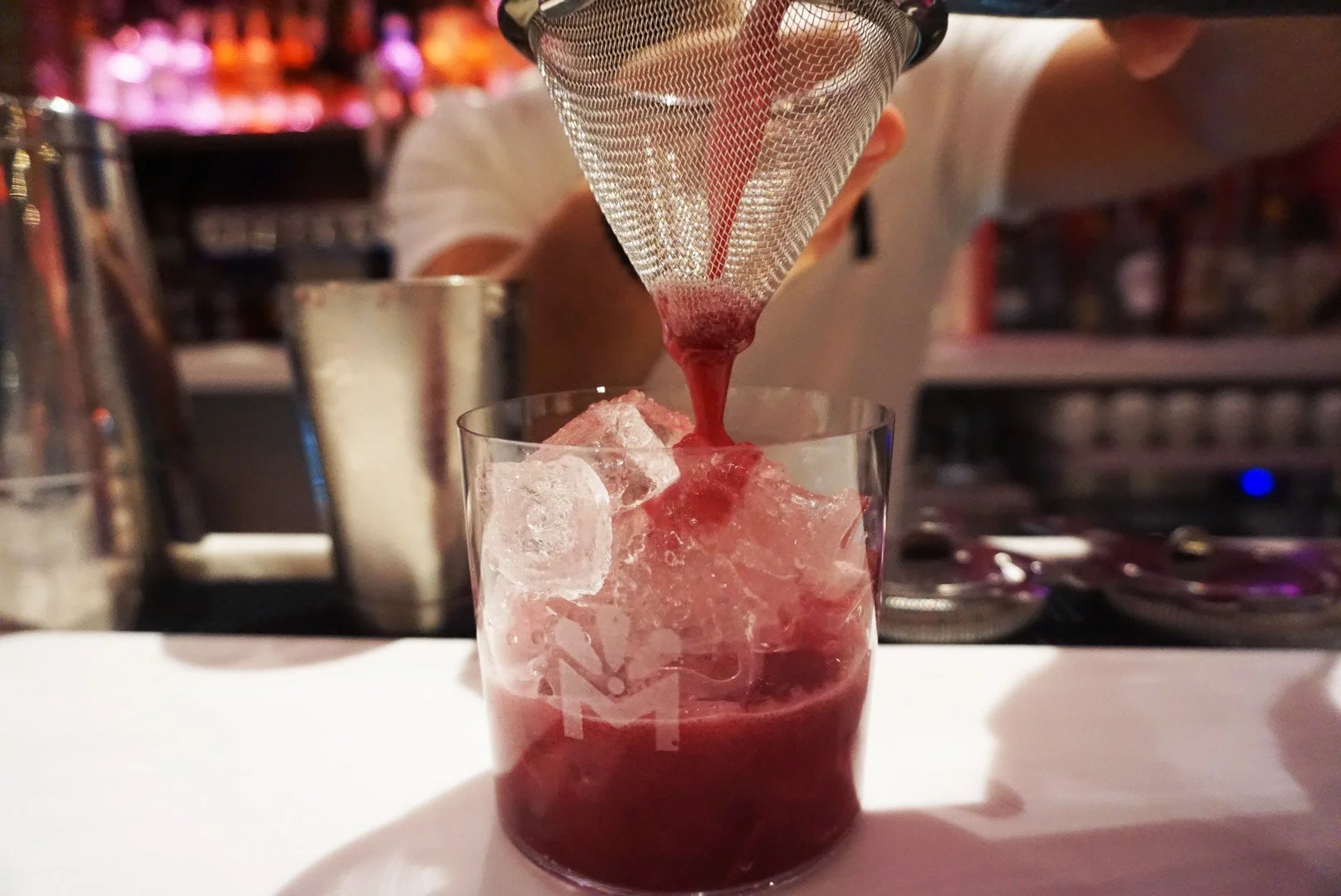 A bartender pours a red liquid through a strainer into a glass filled with ice at a bar.