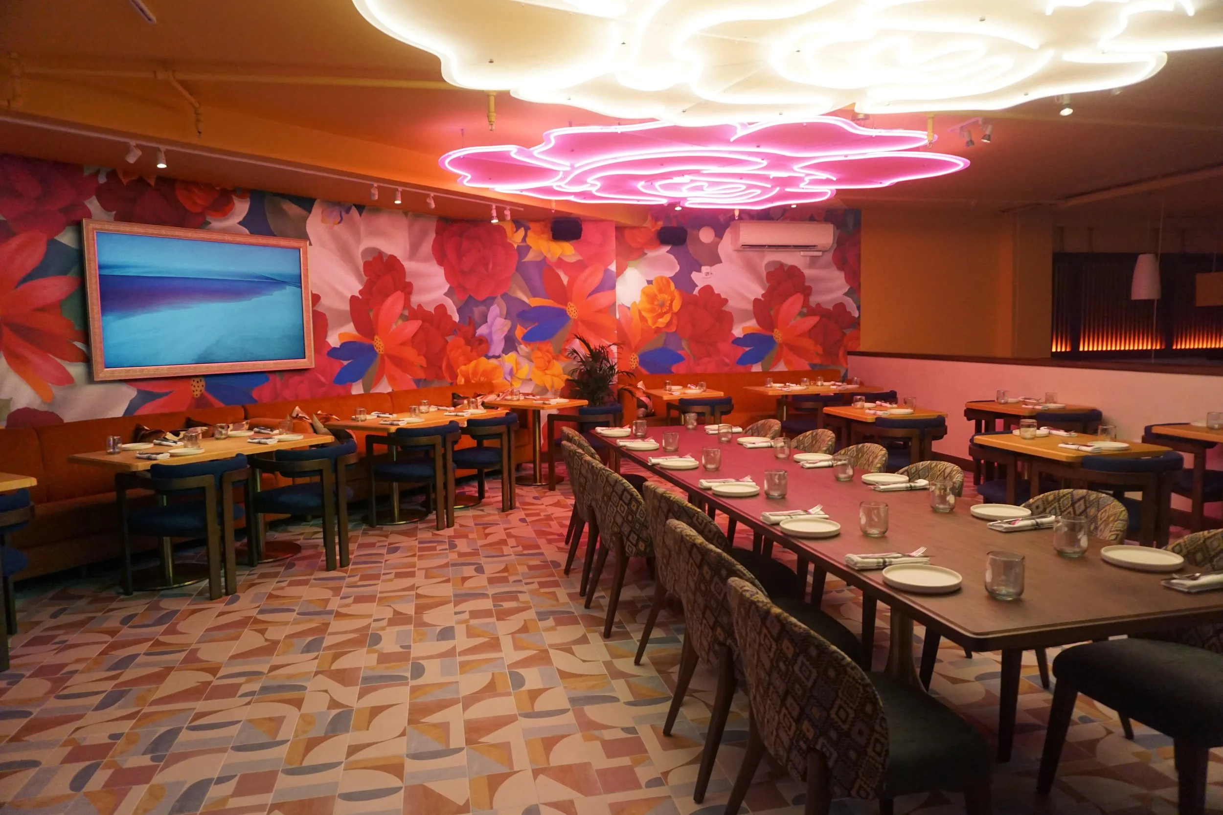 Dining room with modern decor, floral mural wall, pink neon cloud ceiling lights, tables set with plates and glasses, colorful patterned chairs, and a large flat-screen TV on the wall.
