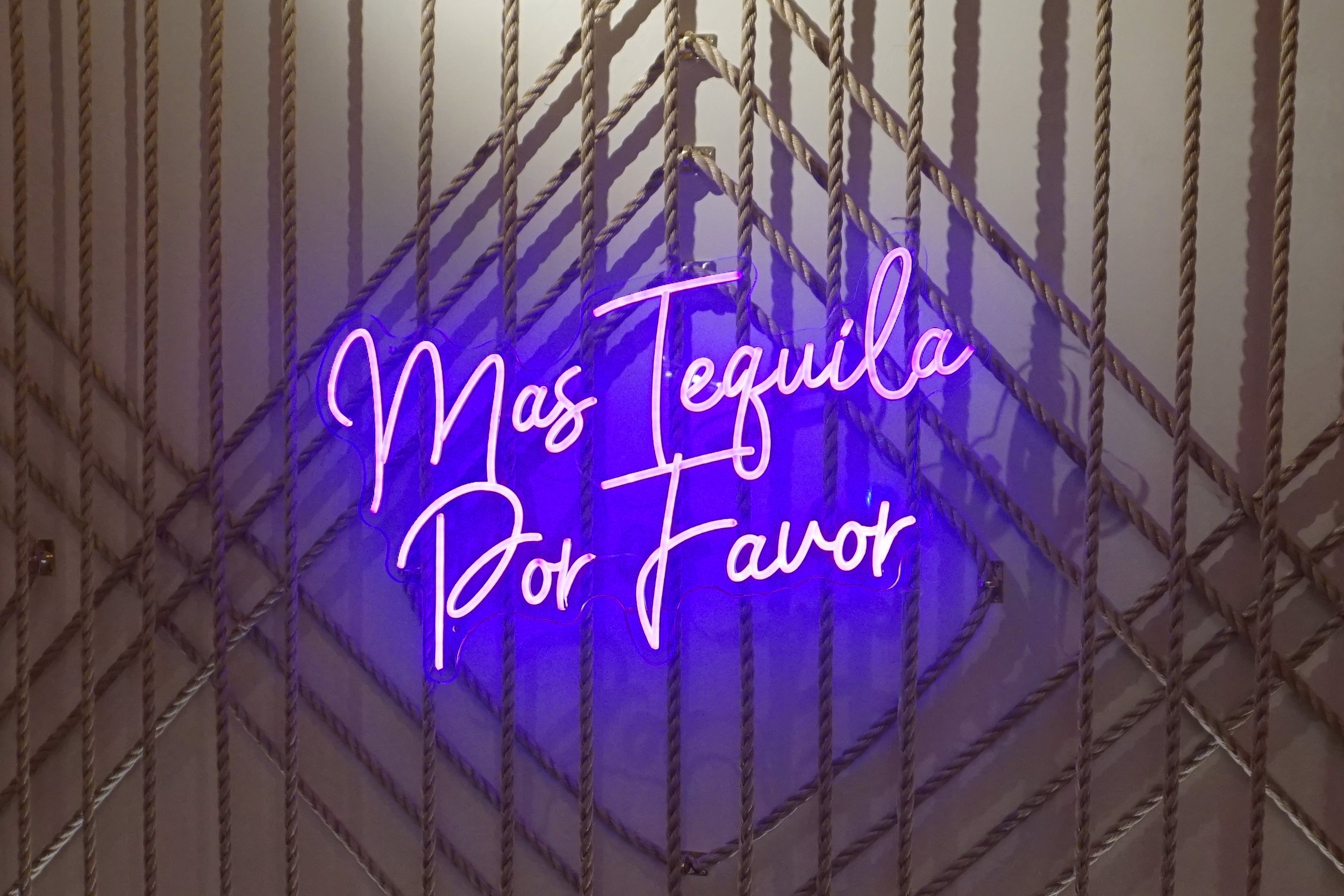 Neon sign reading 'Mas Tequila Por Favor' in pink and purple, mounted on a wall with decorative ropes.