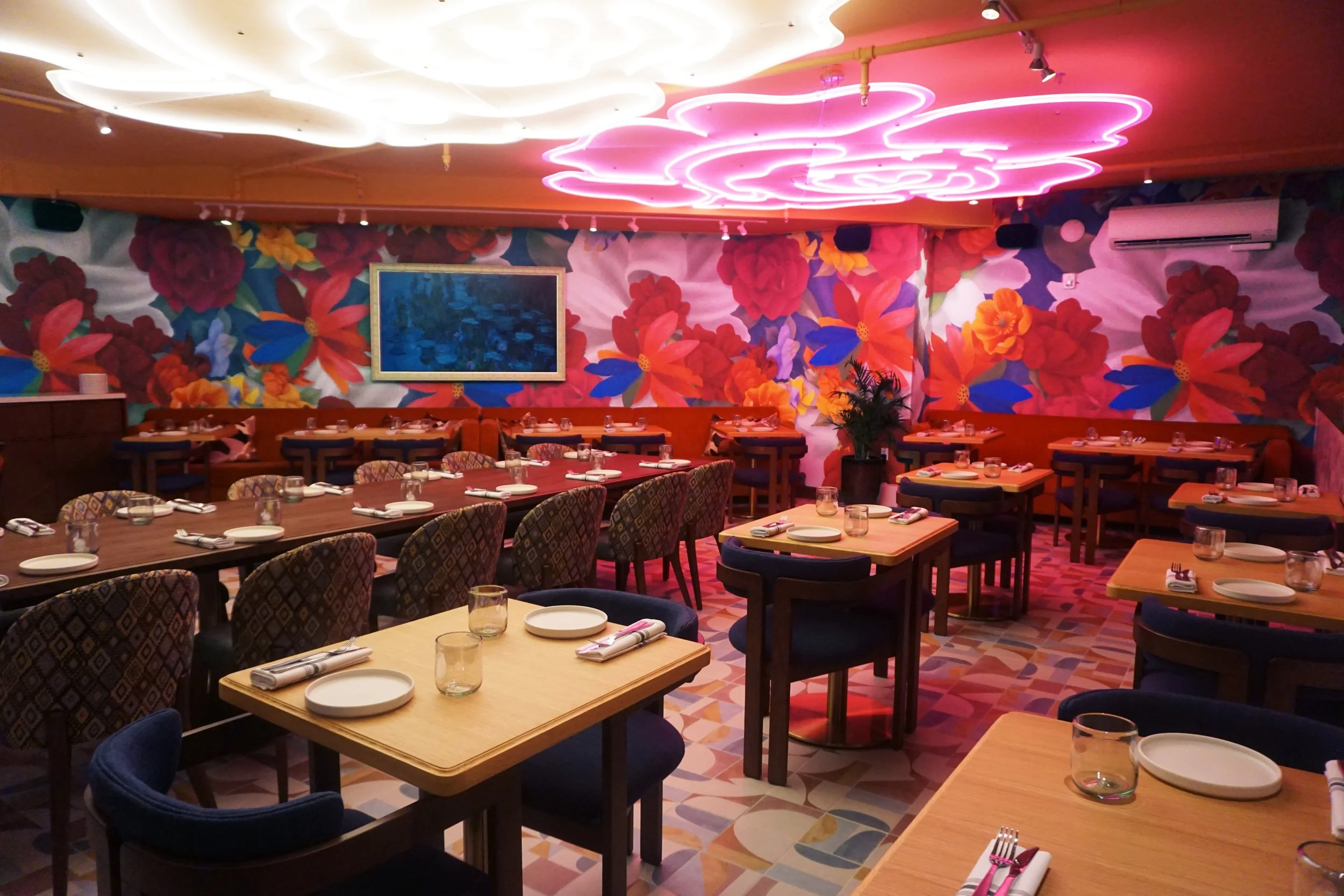 Interior of a restaurant with colorful floral wall mural, tables set with plates, glasses, and utensils, and pink neon cloud-shaped ceiling lights.