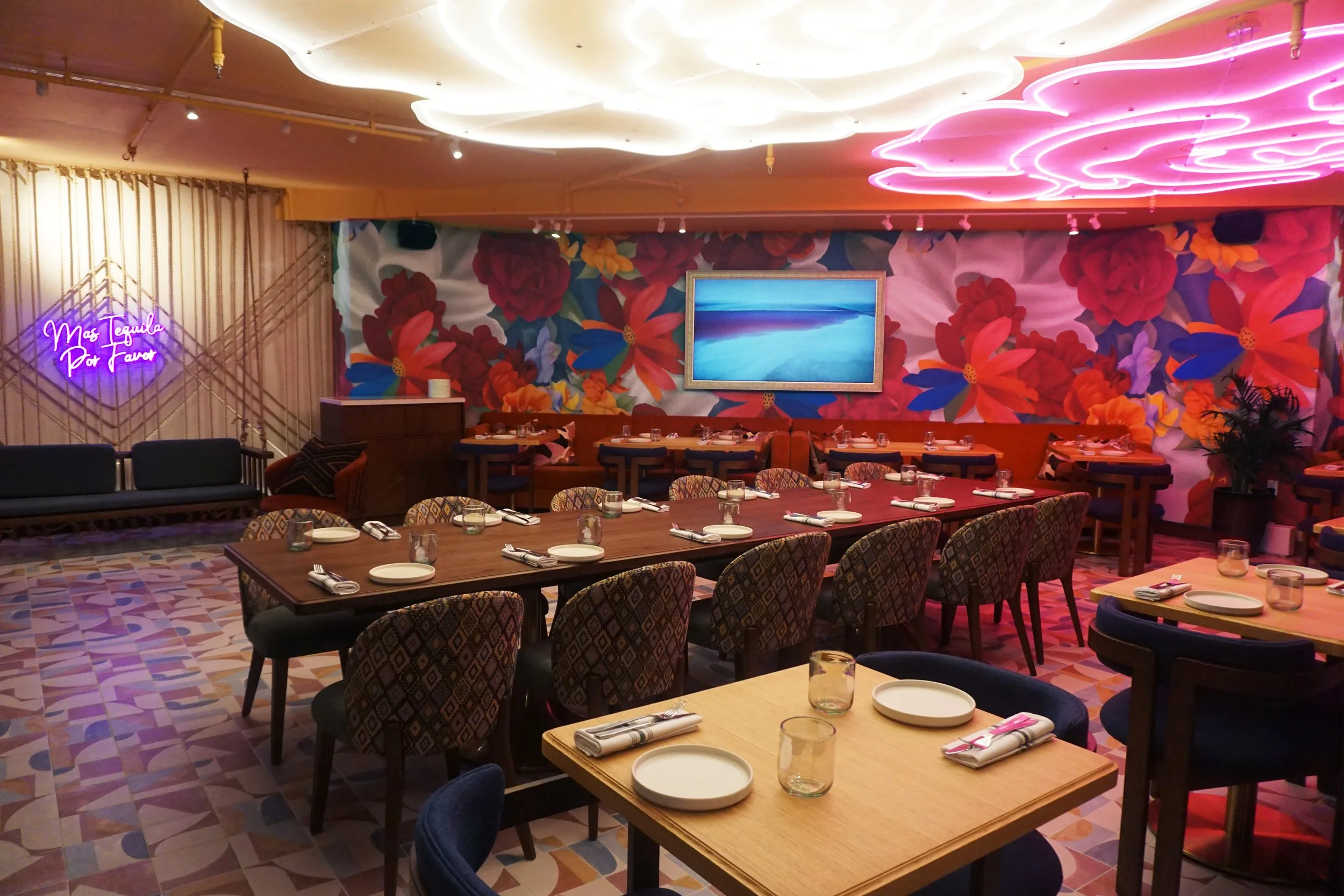 Interior of a restaurant with tables set for dining, a floral mural on the wall, a painting, and a neon sign that says 'Más Tequila Por Favor'