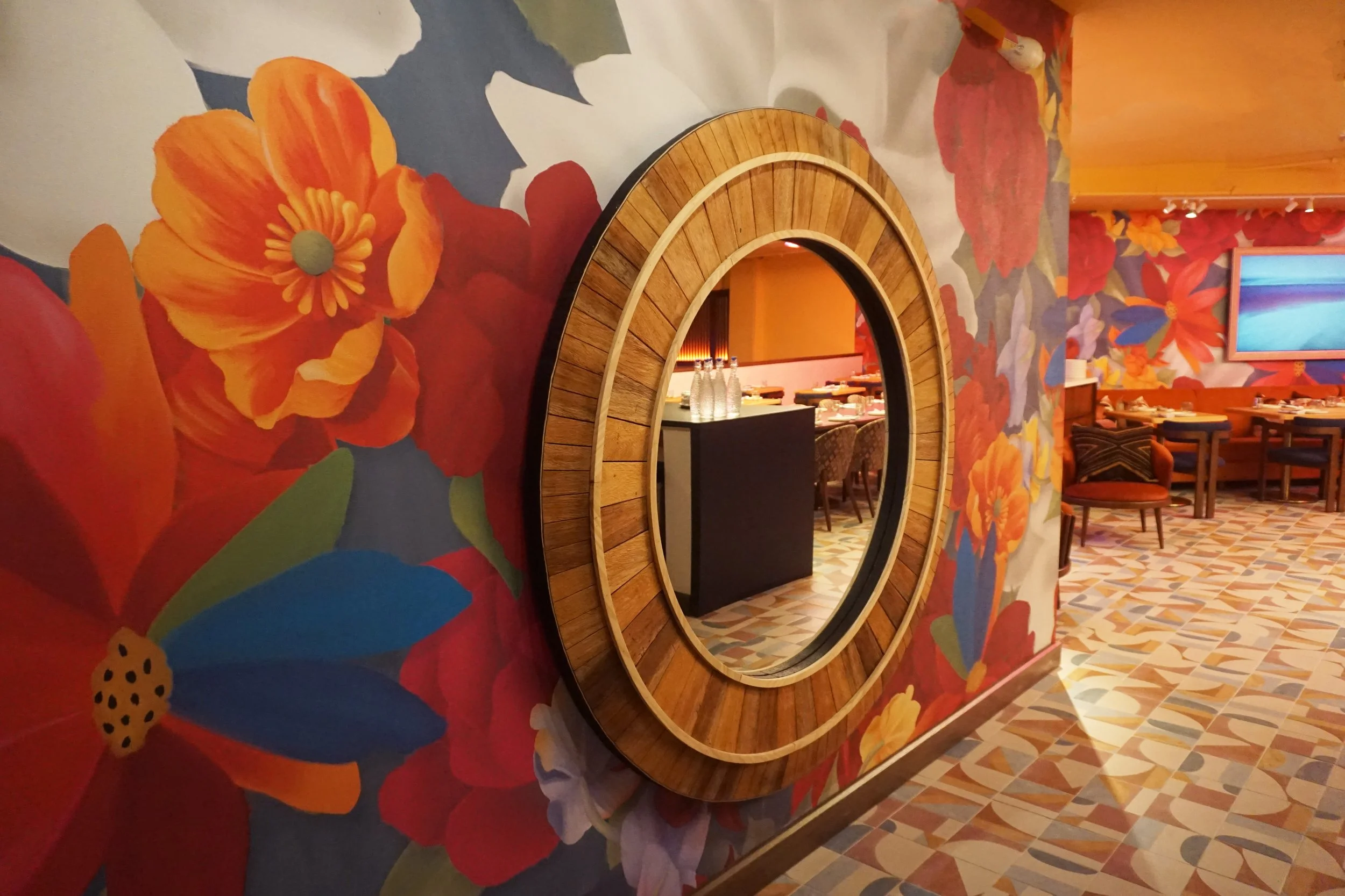 A large round mirror with a wooden frame is mounted on a colorful floral mural wall inside a restaurant. Reflects part of the dining area with tables, bottles of water, and chairs.