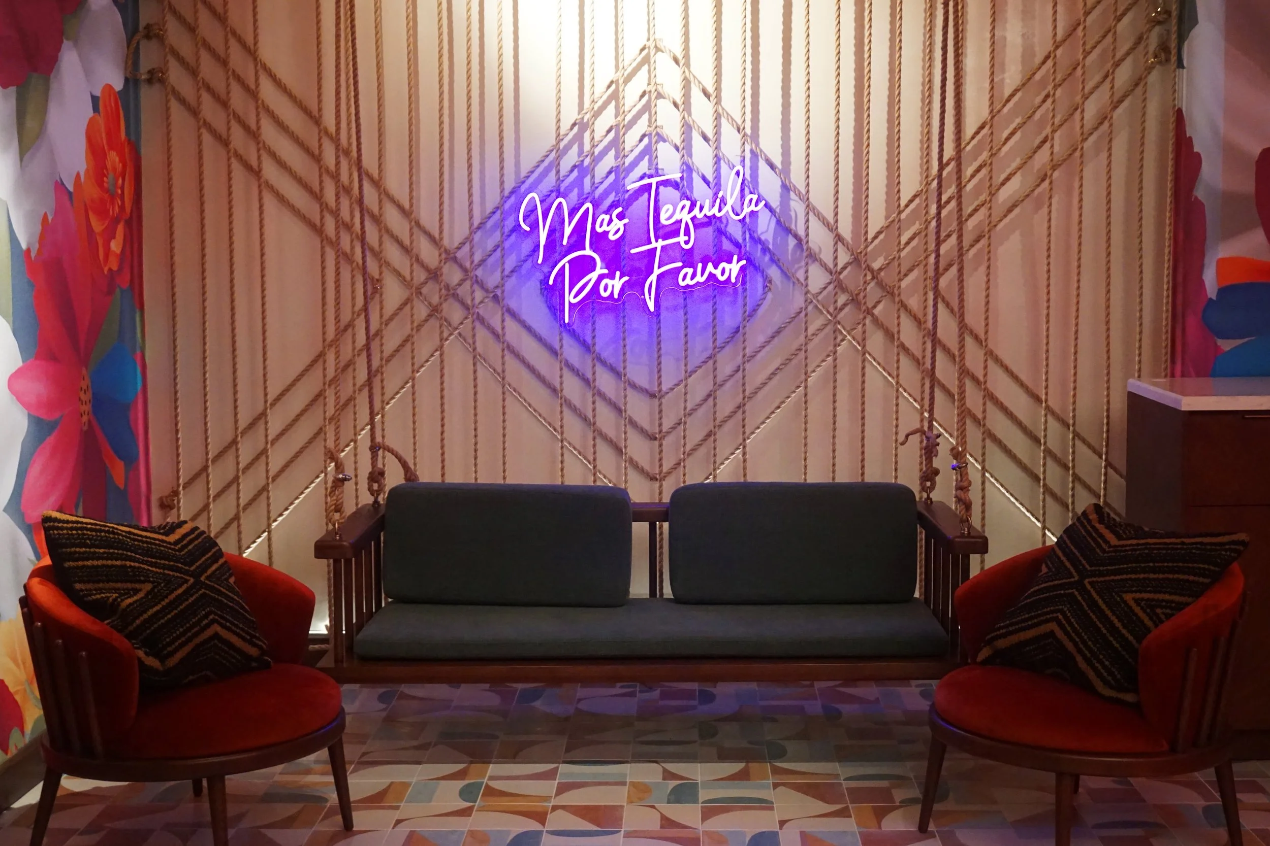A cozy seating area with two red velvet chairs and a dark green couch, decorated with patterned pillows. Behind the seating is a wall with a geometric gold rope design and a neon sign that reads 'Mas Tequila Por Favor' in purple script. The floor has