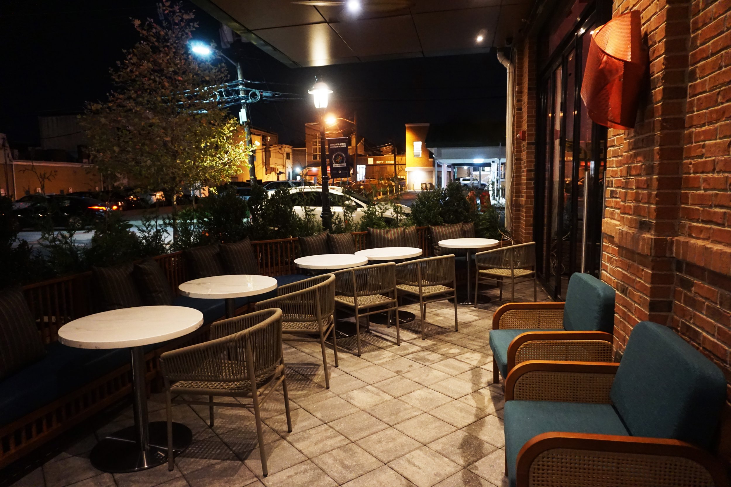 Empty outdoor patio at night with white round tables, blue and rattan chairs, brick wall with a red furnishing, and street view with parked cars and streetlights in the background.