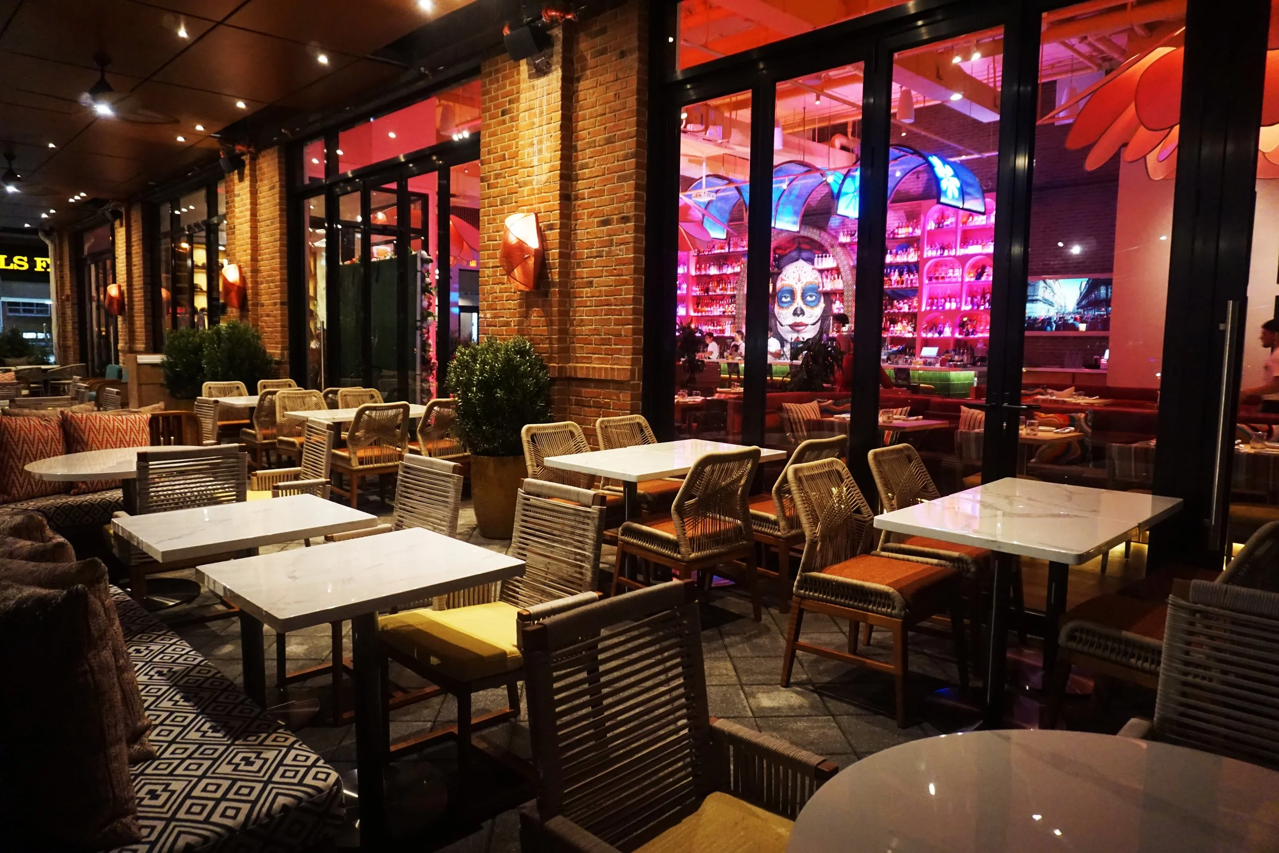 Empty outdoor patio of a restaurant with white marble tables, wicker chairs, and potted plants, with a colorful indoor bar with neon blue and pink lights visible through glass windows.