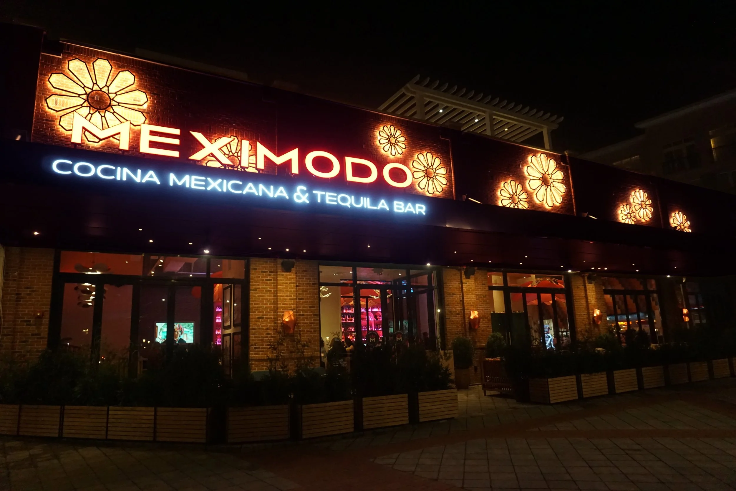 Night view of Meximo restaurant with illuminated sign, brick front, and large windows showing inside dining area, decorated with warm lighting and sombrero shaped light fixtures.