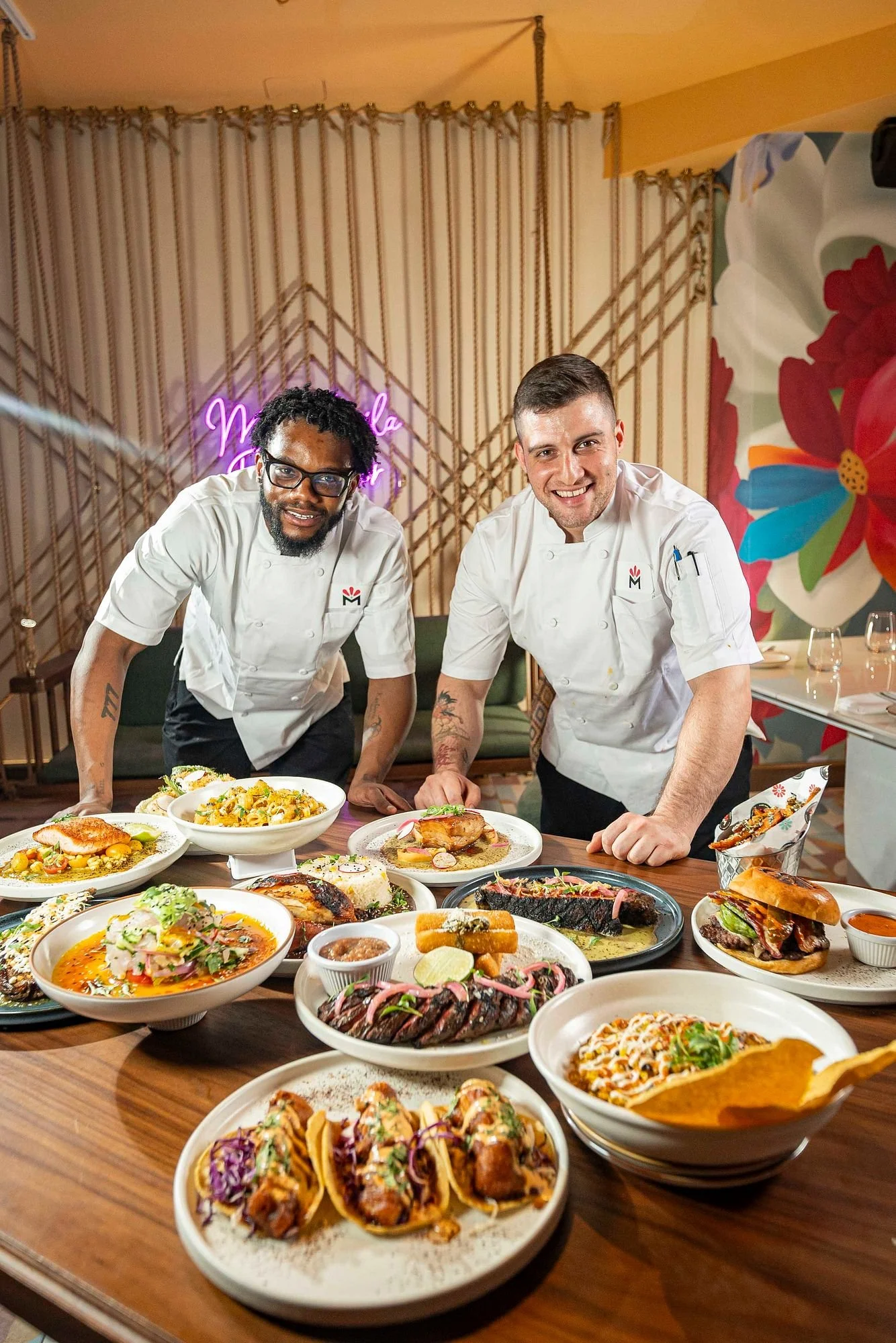 Two chefs standing behind a table filled with various dishes in a restaurant setting, smiling at the camera.