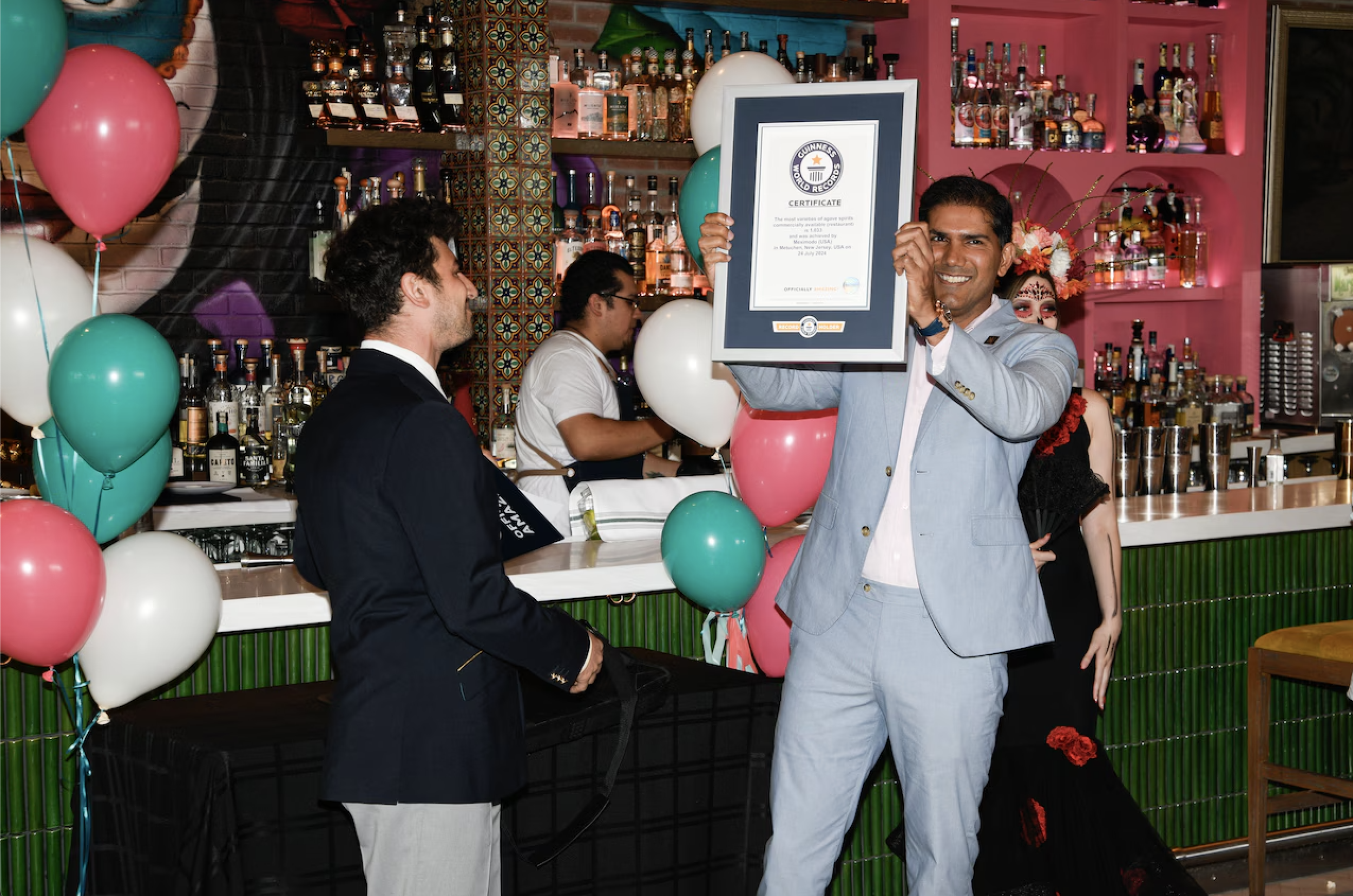 A man in a light gray suit holding a Guinness World Records certificate, smiling at a celebration event with balloons and a decorated bar in the background.