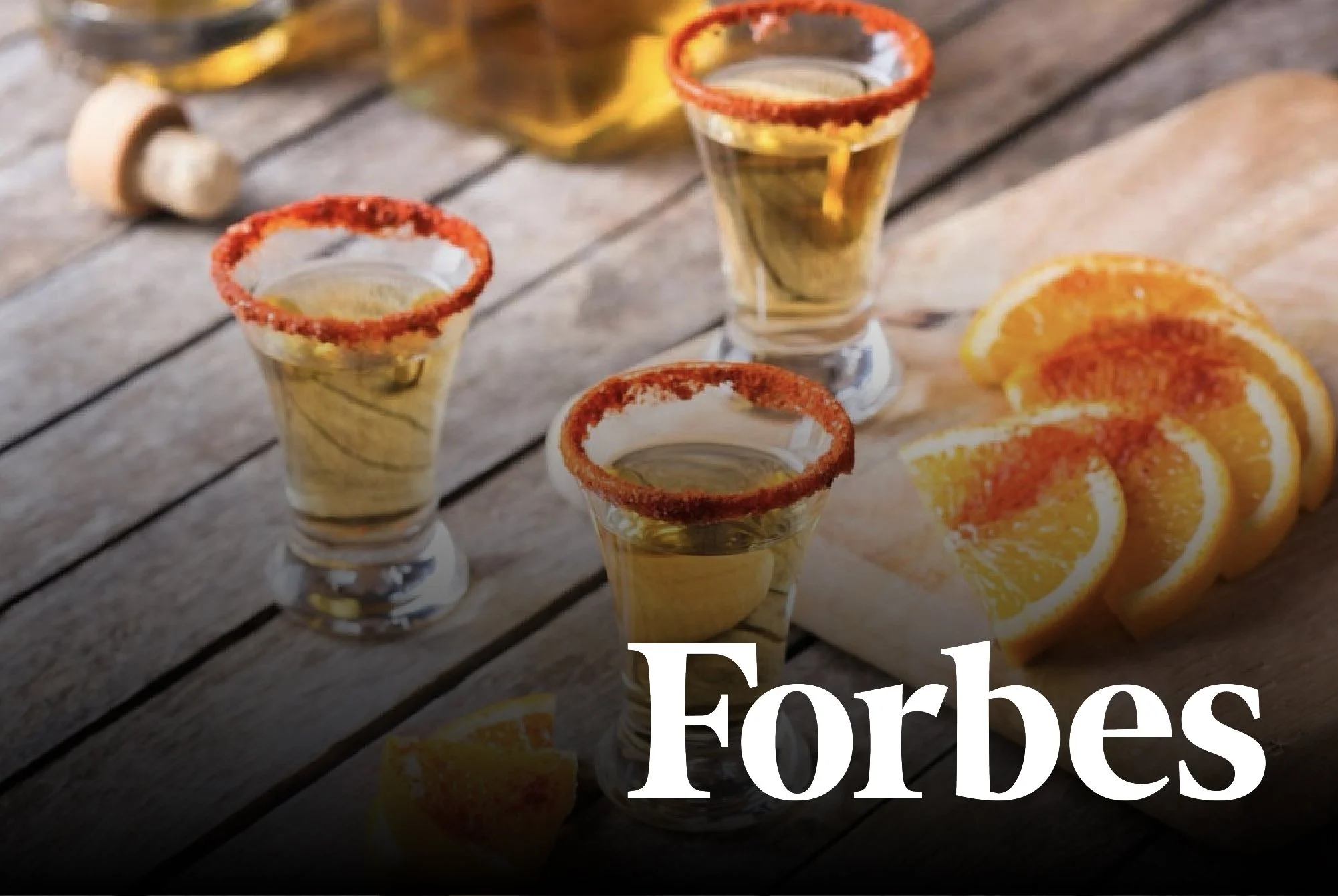 Three shot glasses filled with a yellow beverage, garnished with red chili powder on the rim, placed on a rustic wooden table. To the right, there are several orange slices on a cutting board. In the background, there is a bottle of golden liquor.