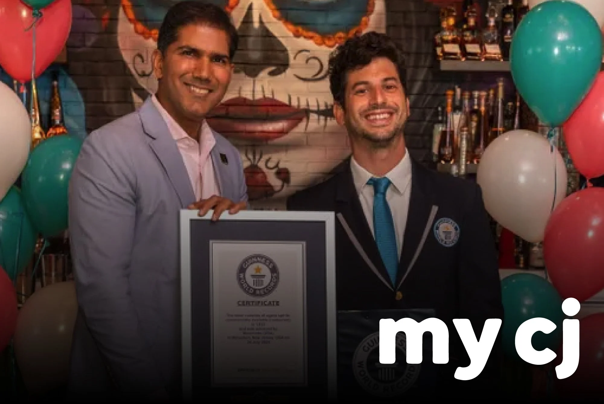 Two men dressed in suits smiling at a celebration, with balloons and a decorated background, one holding a framed certificate.