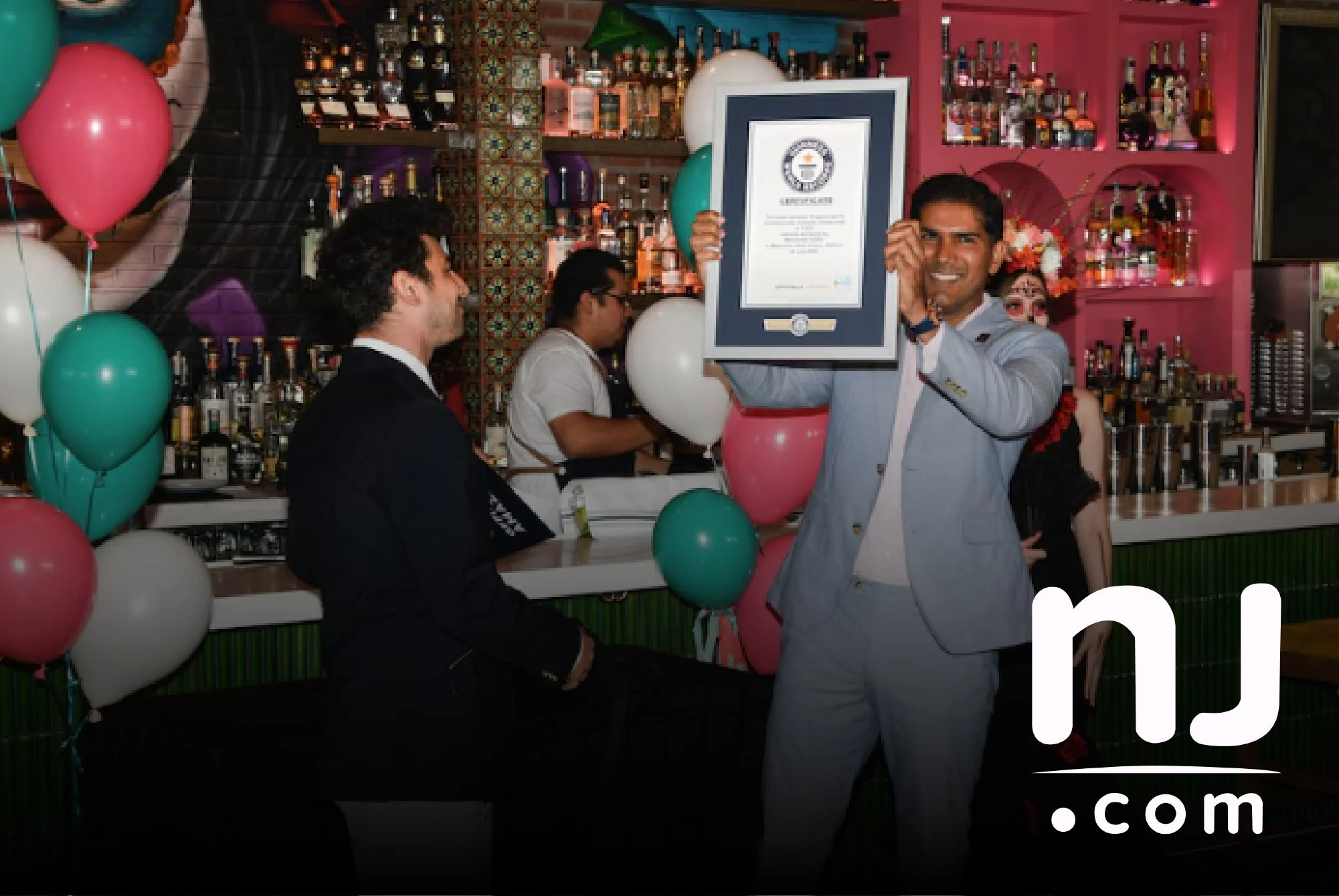 A man in a gray suit holding a framed Guinness World Records certificate and smiling at a celebration with pink and white balloons, in a bar setting with colorful bottles on shelves in the background.