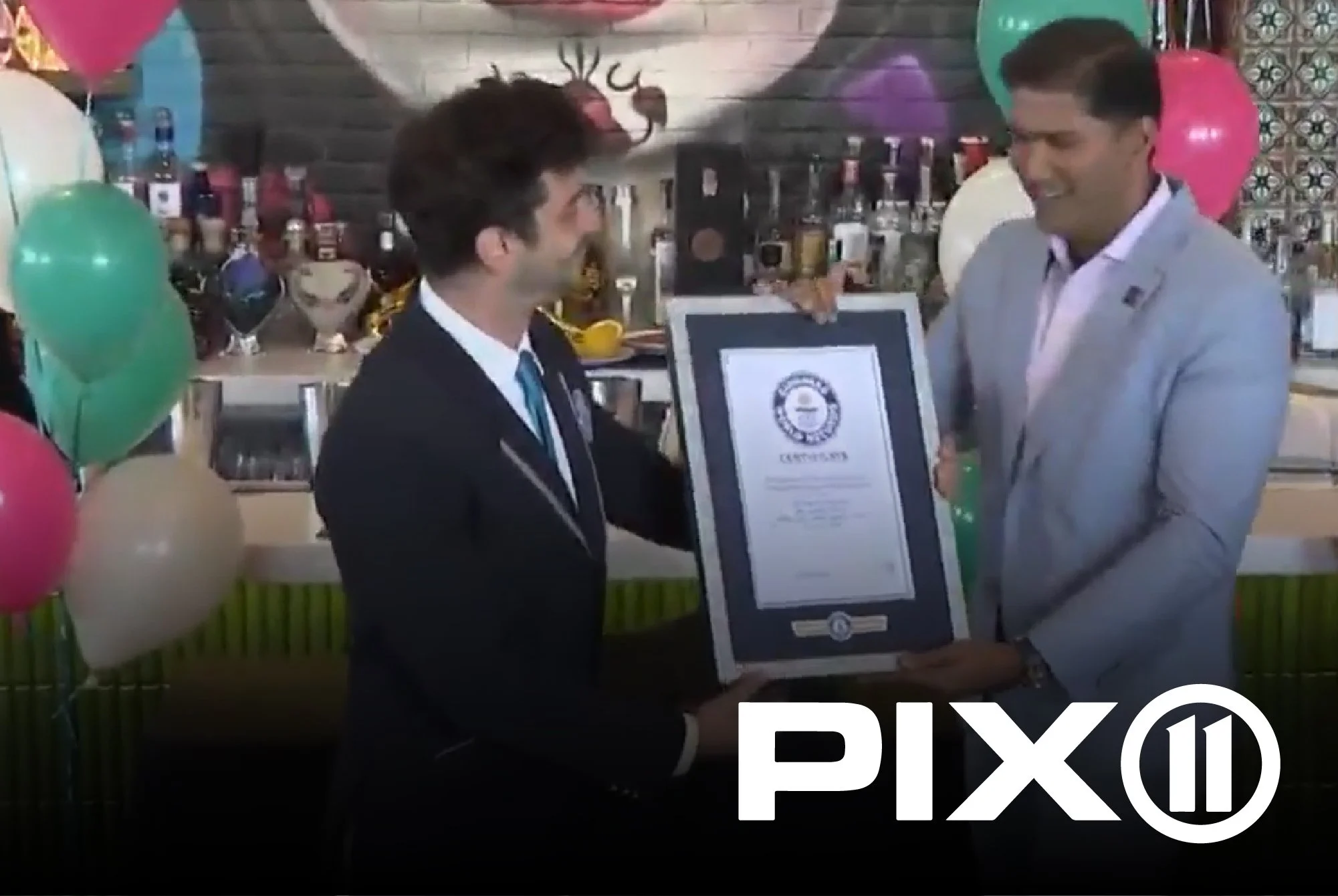 Two men at a celebration holding an award certificate, surrounded by colorful balloons and a bar area in the background.