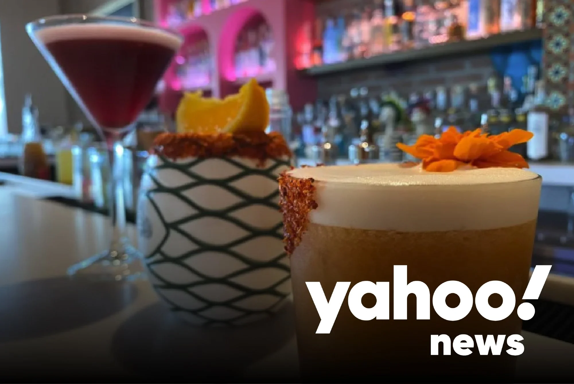 A bar scene with a cocktail on the counter, a basket with a lemon wedge, and a martini cocktail in a martini glass. The background has shelves with bottles and colorful lighting. The image has a 'Yahoo News' logo in the bottom right corner.