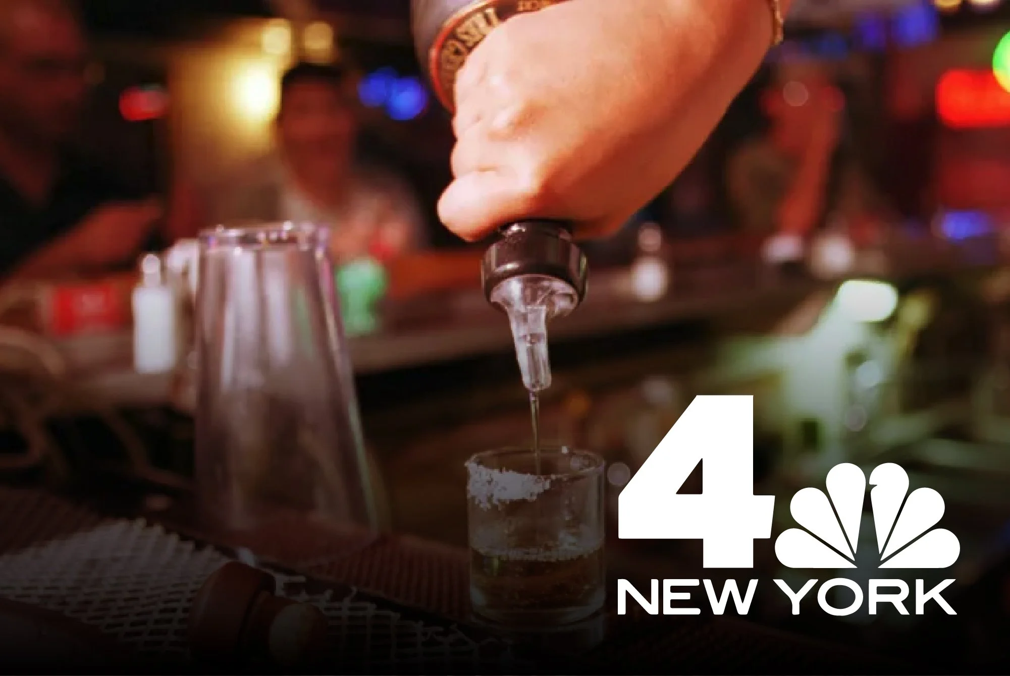 Person pouring a drink from a bottle into a shot glass on a bar counter with blurred people in the background, logo of NBC New York in the corner.