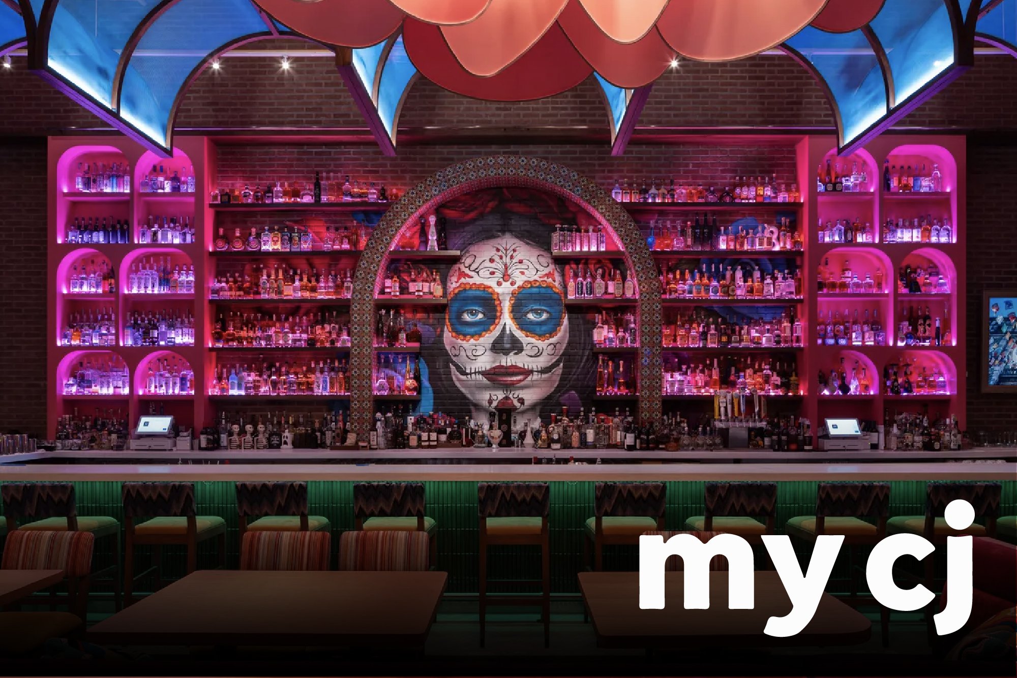 Bar with colorful shelves of liquor bottles, a large painted face of a sugar skull, and pink neon lighting