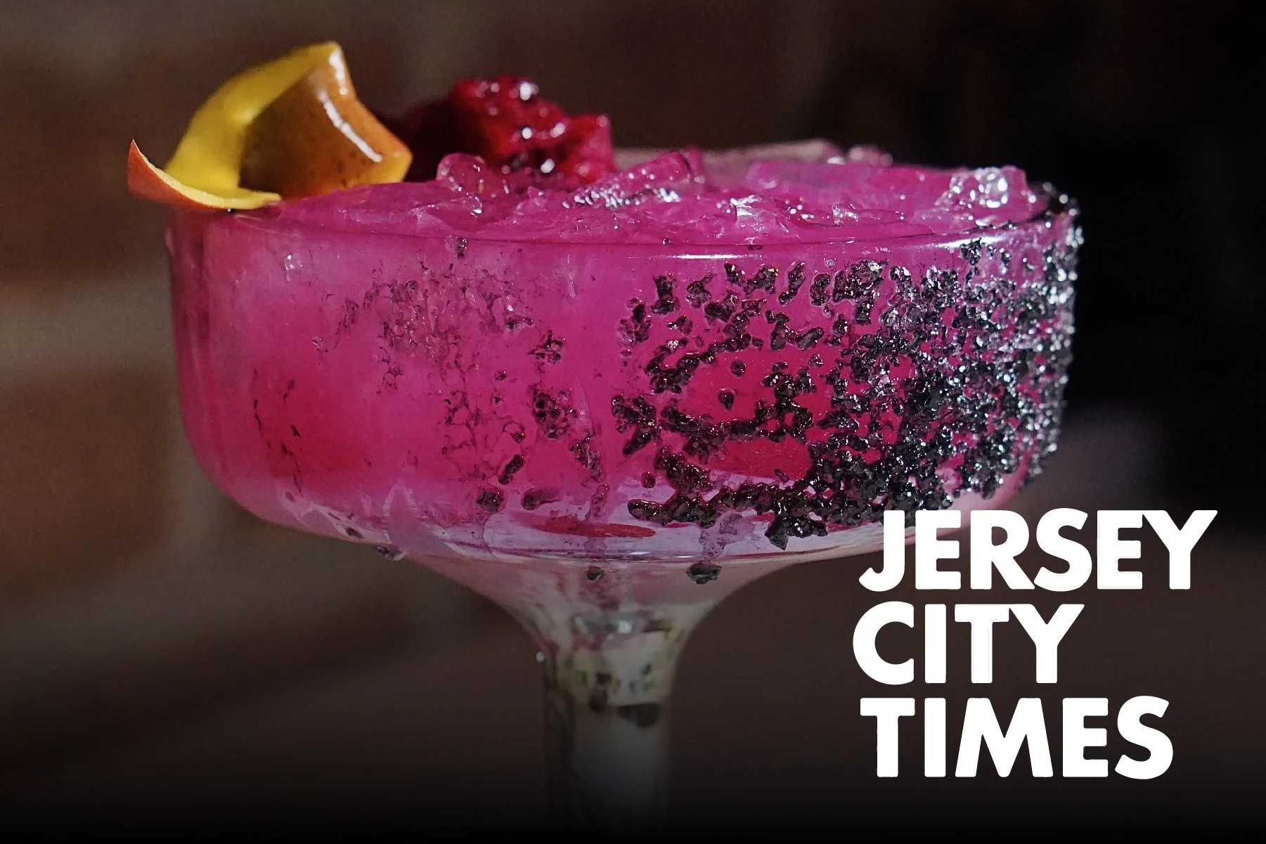 Pink cocktail in a glass with black sugar rim, garnished with a lemon wedge and berries.
