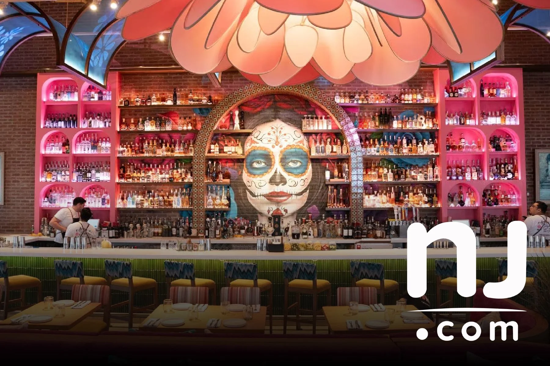 Inside a colorful bar with a large painted sugar skull face on the wall, surrounded by illuminated pink shelves filled with bottles, and a bar counter with bartenders serving drinks. There are tables with chairs in the foreground.
