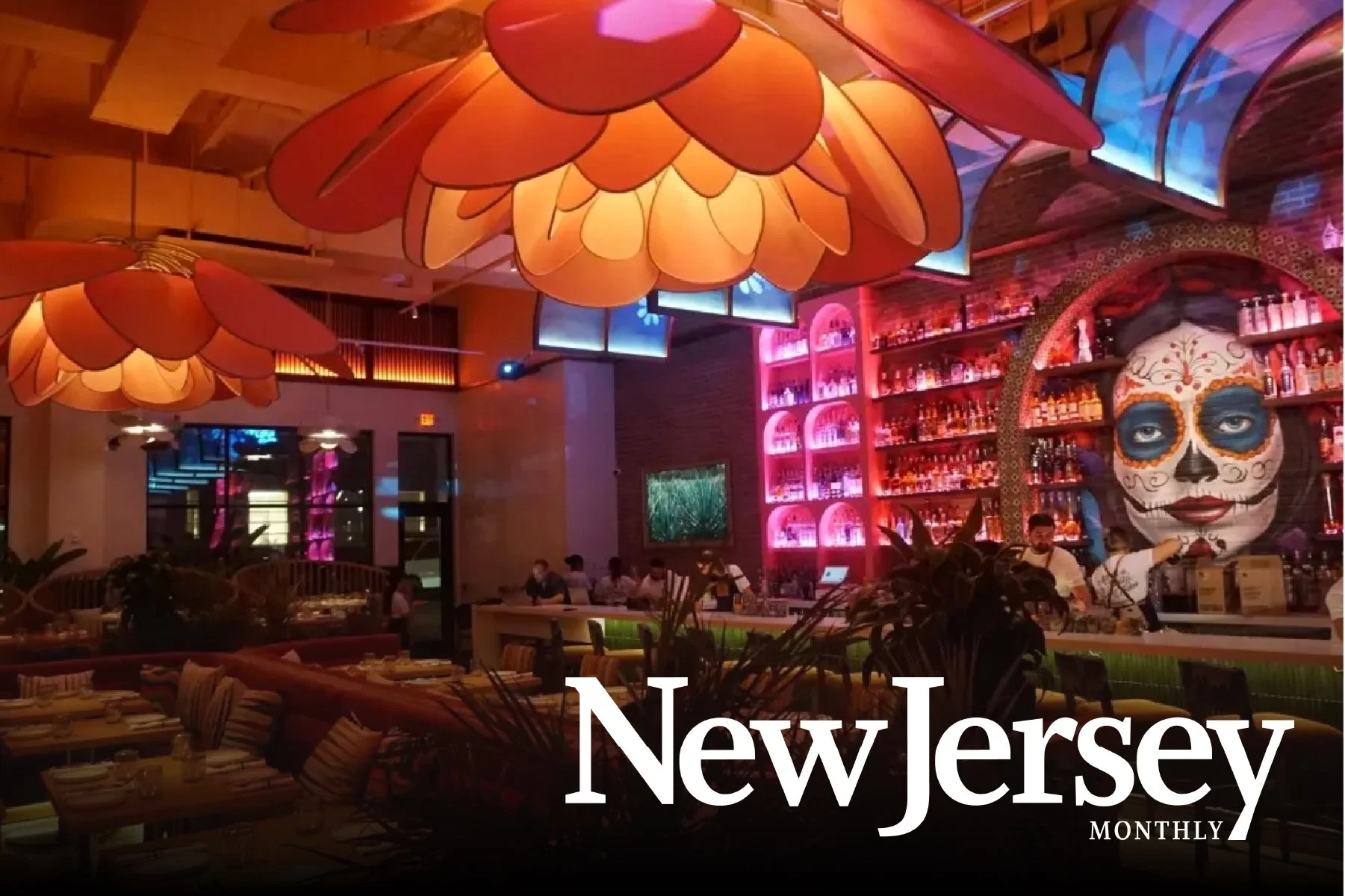Interior of a vibrant restaurant or bar with large flower-shaped pendant lights, a colorful bar area with shelves of bottles, a wall decorated with a large painted sugar skull face, and tables with seating. A few patrons and staff are visible.