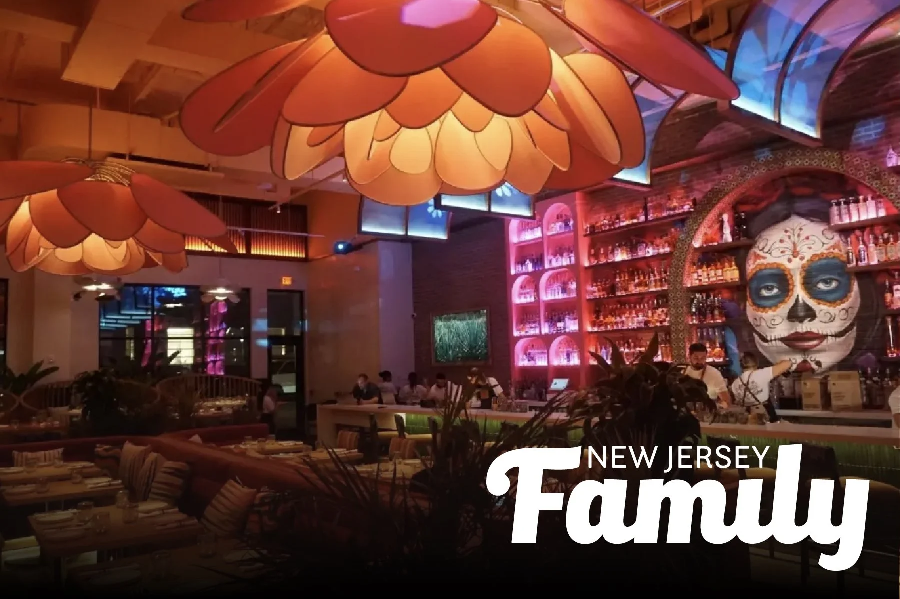 Interior of a lively restaurant or bar with large flower-shaped hanging lamps, a colorful bar area with shelves of bottles, a prominent face painting of a woman with sugar skull makeup, and patrons dining and working at tables.