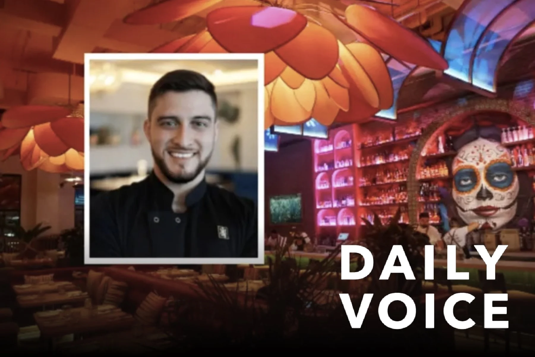 A smiling man in a chef's uniform with a black jacket, standing inside a busy restaurant. The restaurant has colorful, festive decorations, including a large sugar skull face painting and vibrant lighting, suggesting a themed celebration or Día de los Muertos.