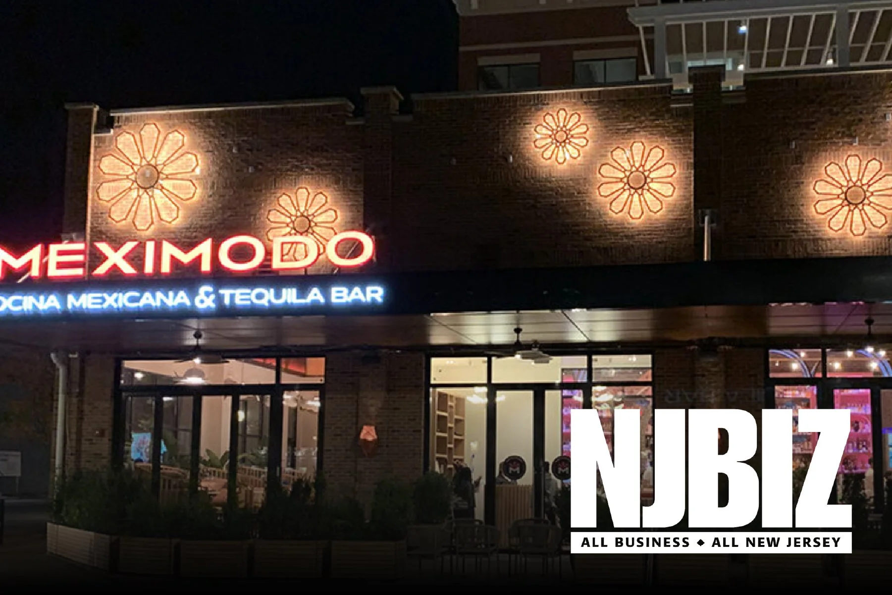 Nighttime exterior view of Meximodo, a Mexican restaurant and tequila bar, with illuminated signage and decorative wall lights shaped like flowers.