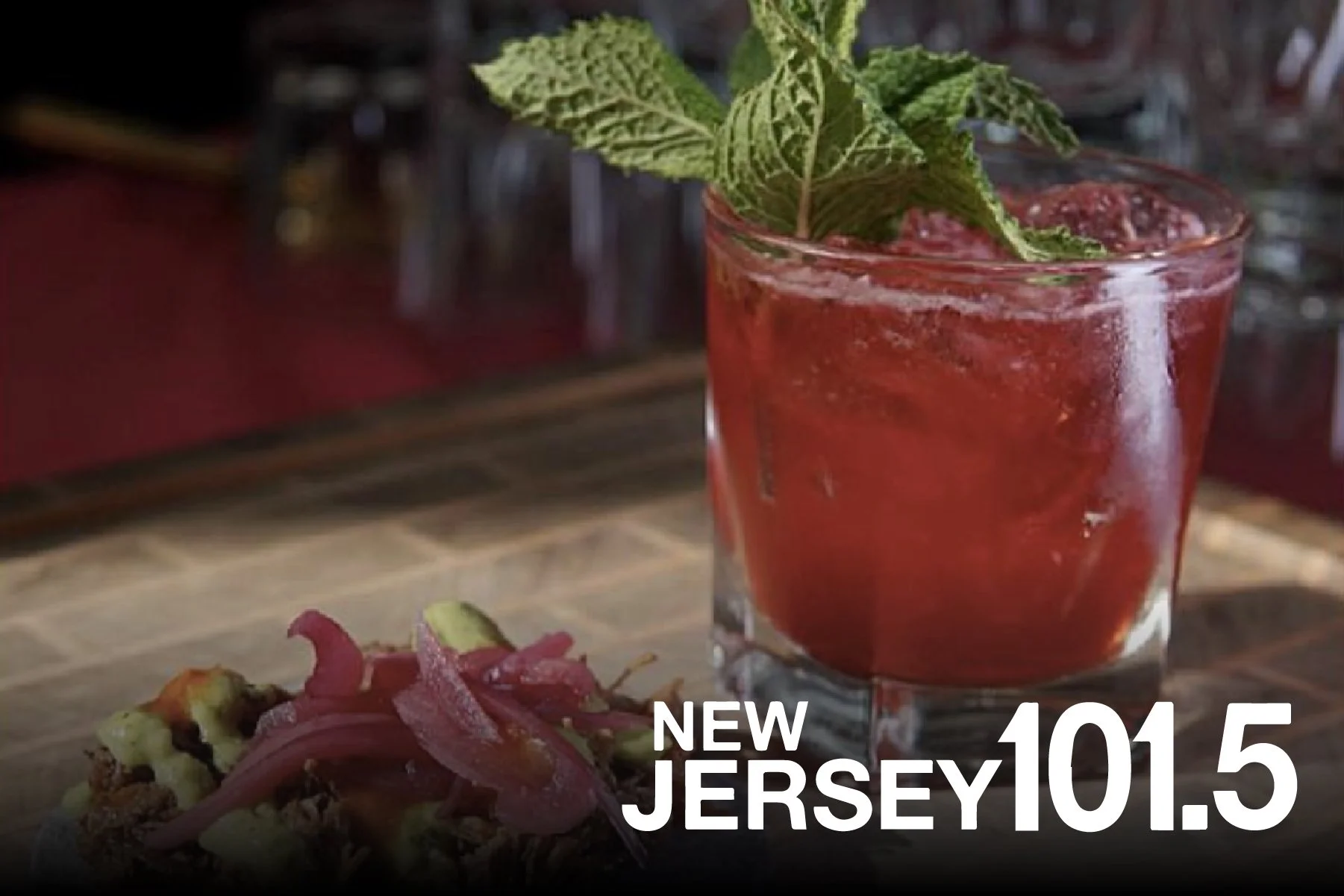 A red cocktail garnished with fresh mint leaves in a clear glass, with a small plate of pickled vegetables on a wooden surface in the foreground. Text overlay says 'New Jersey 101.5'.