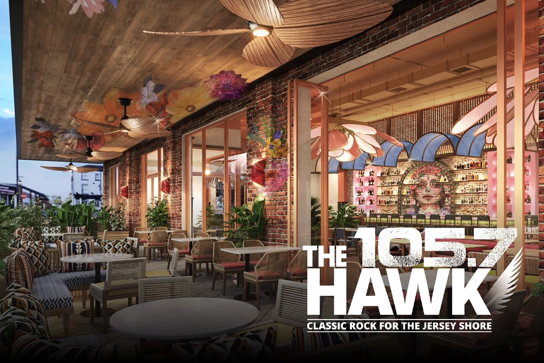 A vibrant outdoor patio area of a restaurant or bar with brick walls, large windows, and lush green plants, featuring ceiling fans, colorful flower decor, and a colorful mural of a woman's face made of bottles on the interior wall, with a logo reading 'The 105.7 Hawk' and the tagline 'Classic Rock for the Jersey Shore'.