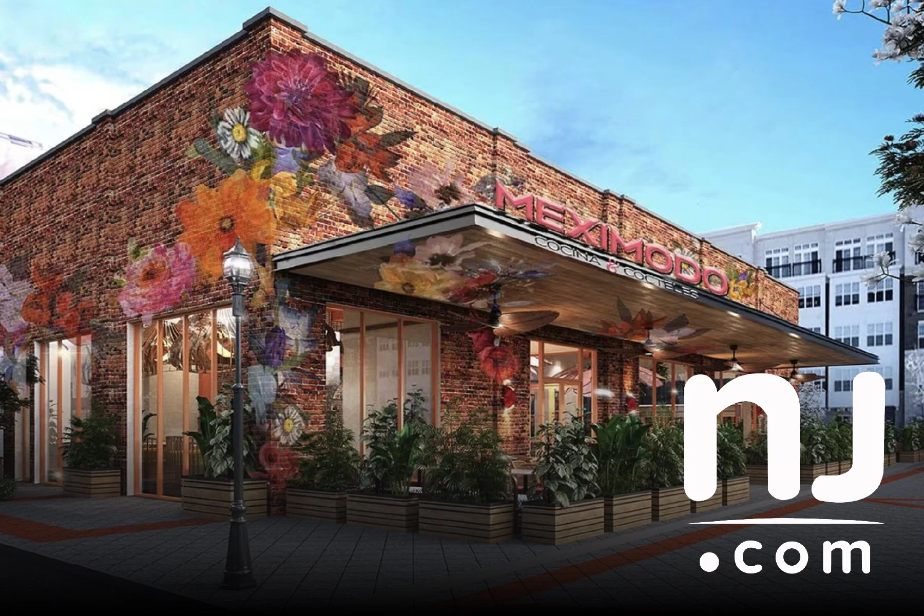 A brick building with large windows and a sign that reads 'Meximodo Cocina & Cocteles,' decorated with a floral mural and surrounded by potted plants, situated in an urban area with clear blue skies.