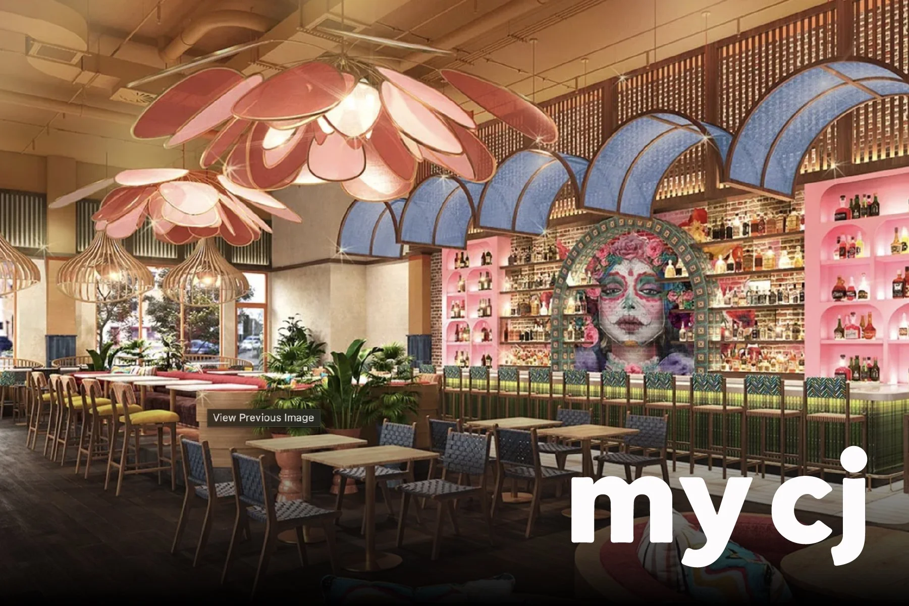 Colorful restaurant interior with pink flower-shaped pendant lights, indoor plants, and a bar area with a mural of a woman's face decorated with flowers, shelves stocked with bottles, and a wooden dining area with chairs and tables.
