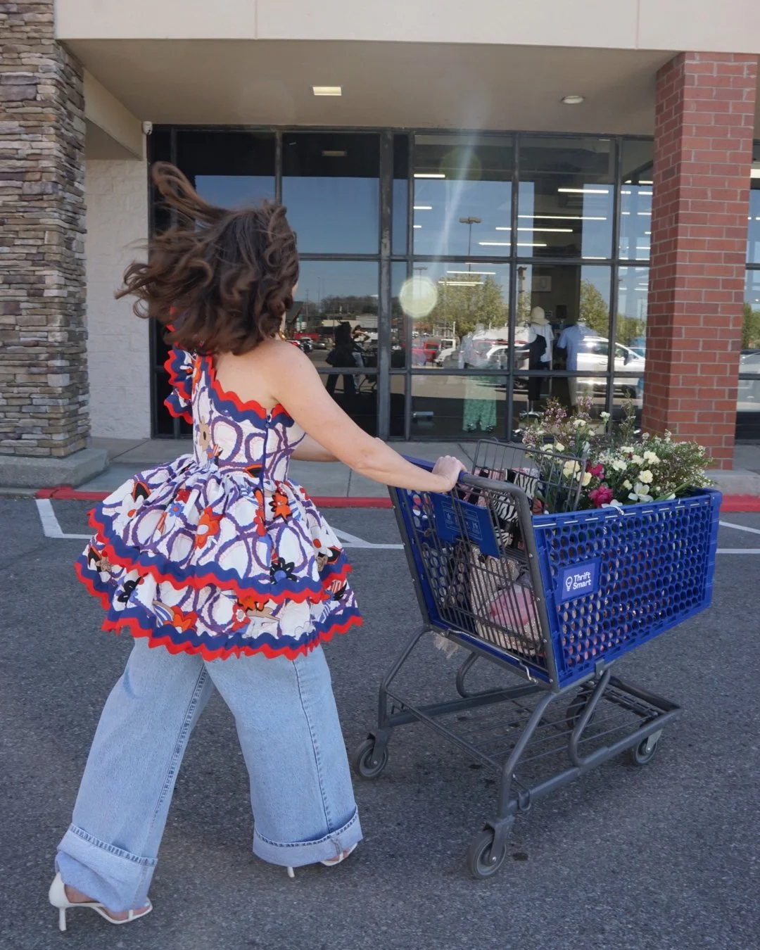 Woman pushing a ThriftSmart shopping cart filled with flowers in a sunny parking lot, wearing a Celia B Anthropologie dress. Sustainable fashion shopper supporting local Nashville thrift store.