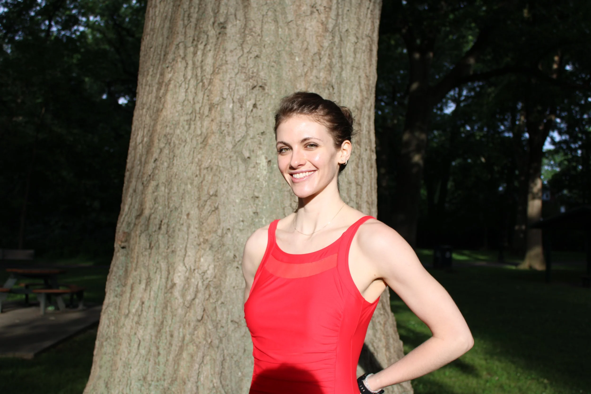 A young woman with short brown hair, wearing a red sleeveless swimsuit, standing outdoors next to a large tree in a park, smiling at the camera.