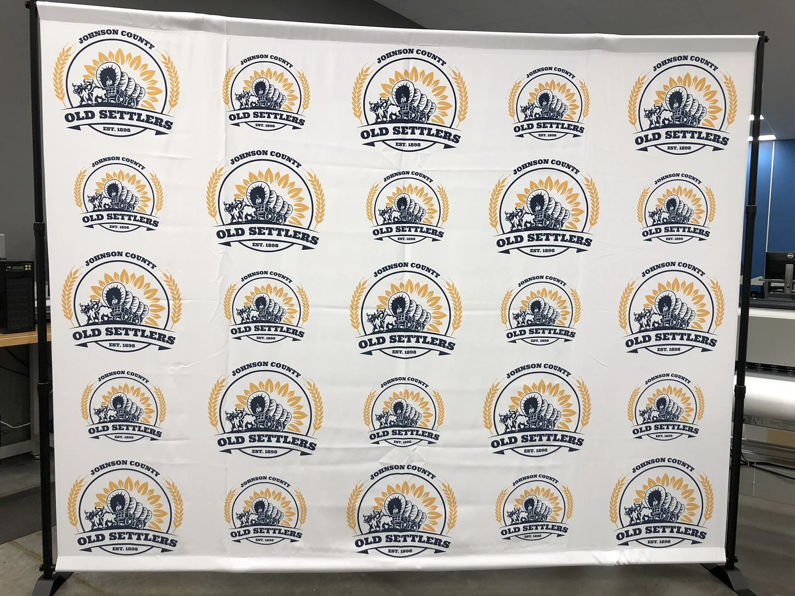 A fabric backdrop with a repeated logo pattern reading 'Johnson County Old Settlers Est 1898', featuring a covered wagon, a horse, and laurels in navy blue, gold, and white.