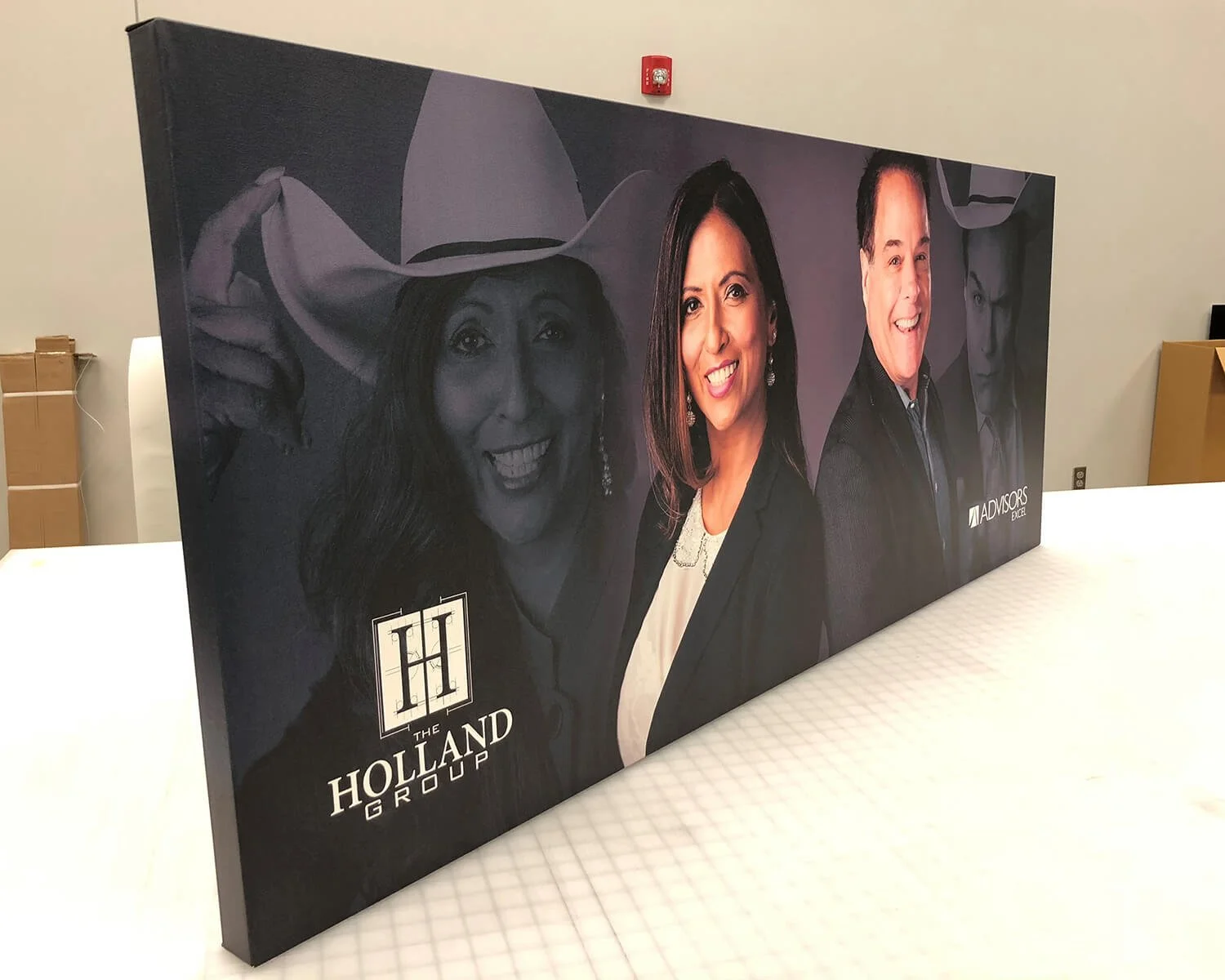 A promotional display with three people, one woman wearing a hat and smiling, the second woman with shoulder-length hair smiling, and a man in a suit, with the logo for The Holland Group and the Advisors Exchange.