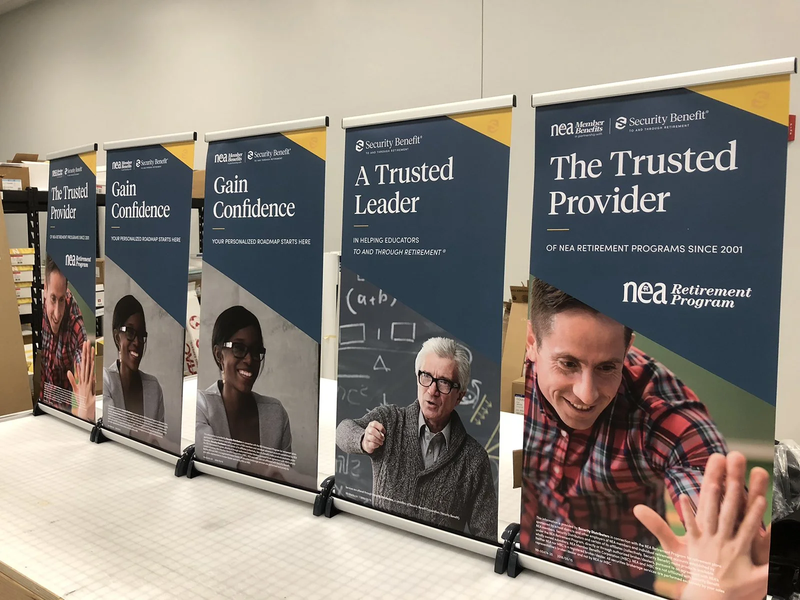 A row of five standing banners for NEA Retirement Program, featuring images of diverse people and slogans like 'Gain Confidence', 'A Trusted Leader', and 'The Trusted Provider'.