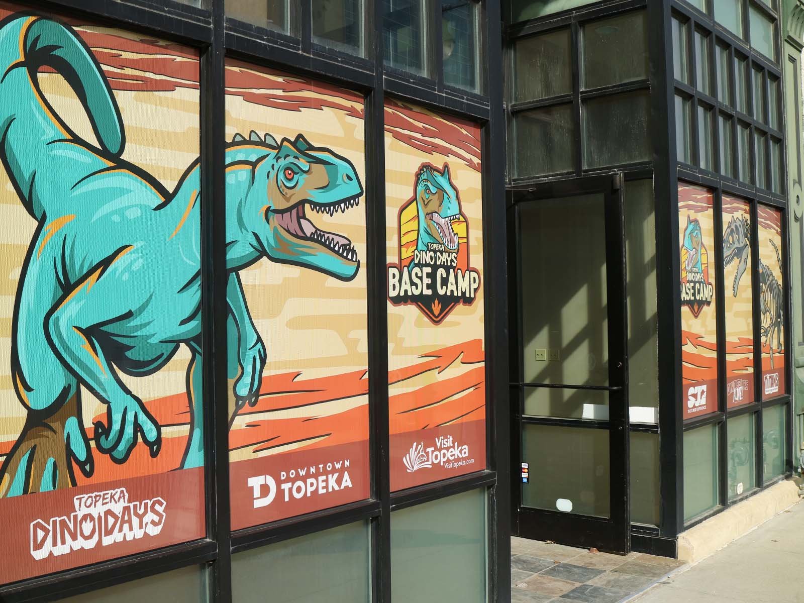 Storefront window with dinosaur-themed promotional posters for Topeka Dino Days Base Camp, featuring a large blue T-Rex and text including "Downtown Topeka" and "Visit Topeka."