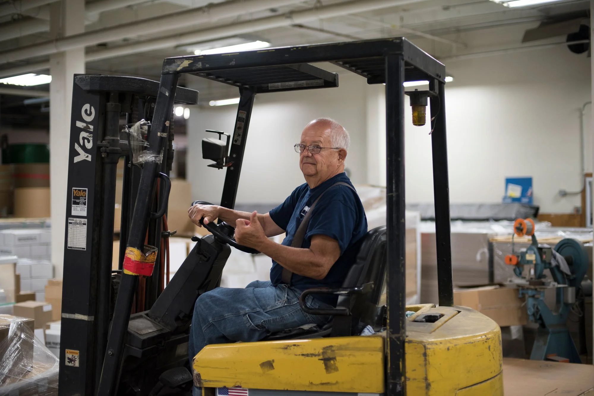 An elderly man driving a yellow Yale forklift inside a warehouse.