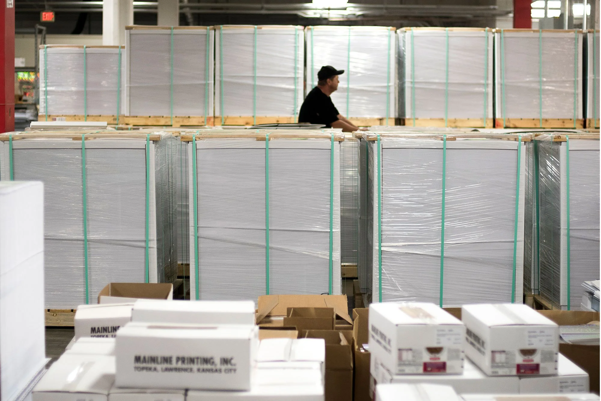 A worker in a warehouse standing behind stacks of large wrapped pallets, surrounded by boxes and storage equipment.
