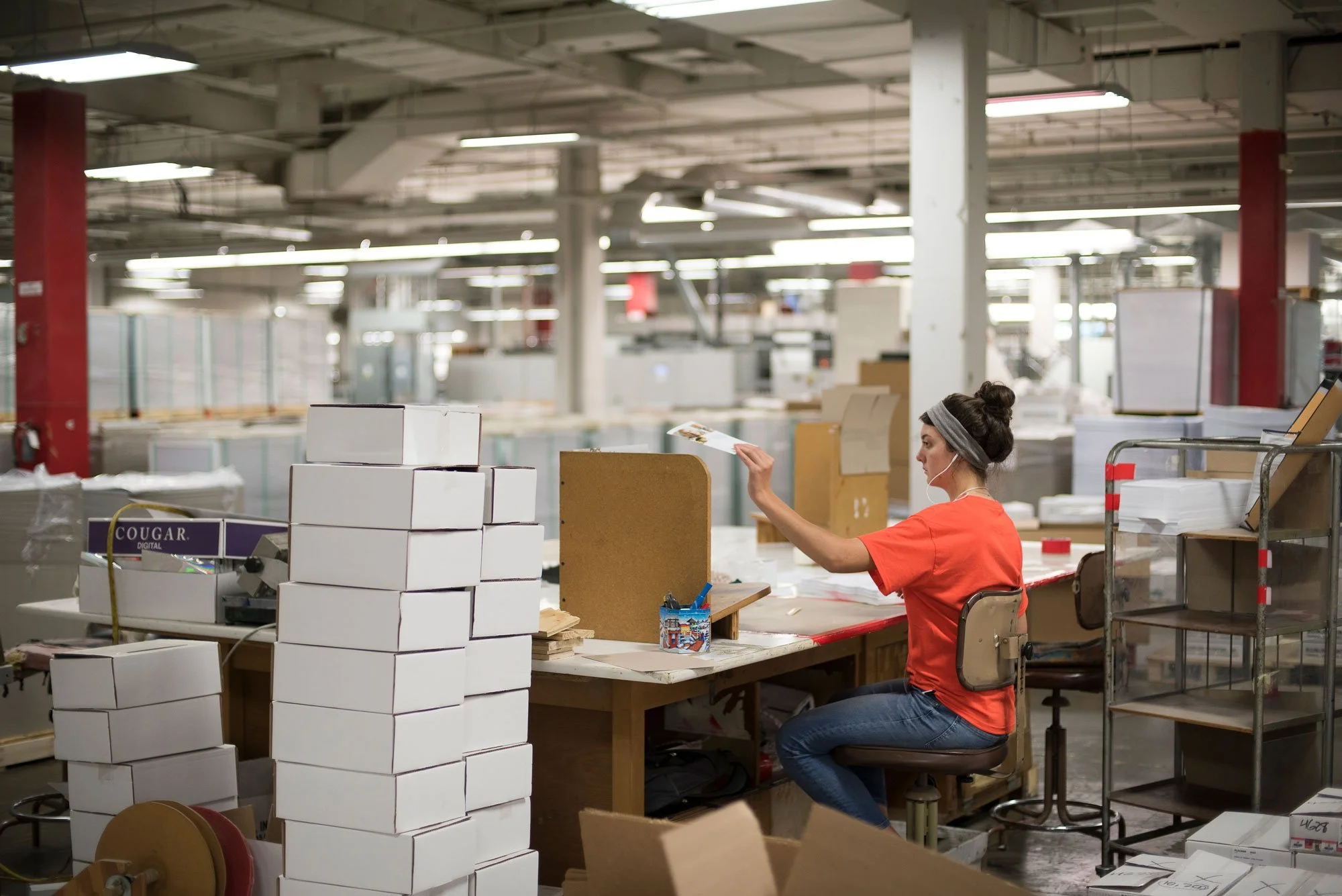 A woman sitting at a worktable in a warehouse or factory, surrounded by stacks of white boxes, working with papers and materials.