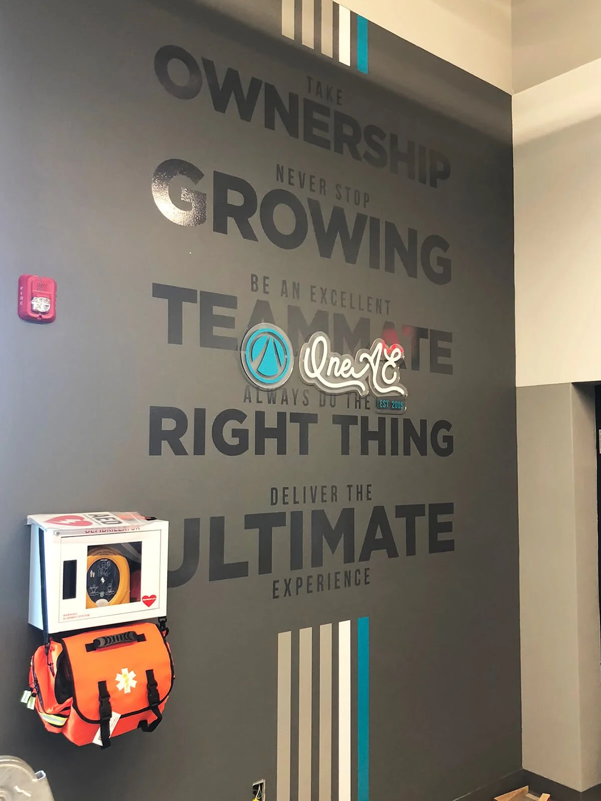 Motivational wall art with text about taking ownership, growth, teamwork, and delivering the ultimate experience, featuring a logo for 'OneXT' and a fire safety kit on the wall.
