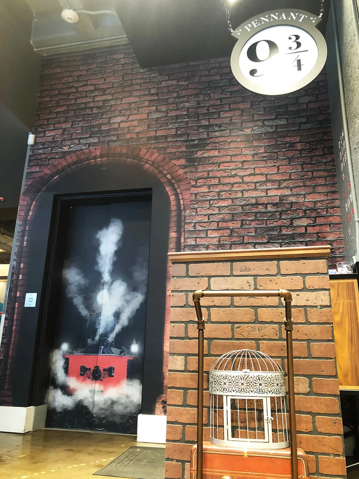Decorative wall with brick pattern, a painted black door with smoke and steam effects, a circular sign reading 'Pennant 9 3 4,' and a white birdcage on an orange container.