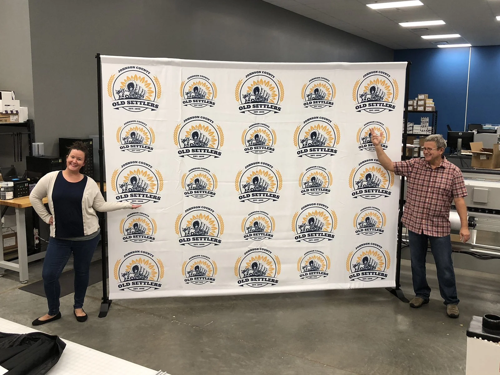 Two people standing in front of a step and repeat banner with logos for Old Settlers and Johnson County, featuring a turkey and wheat design, inside a room with office equipment.