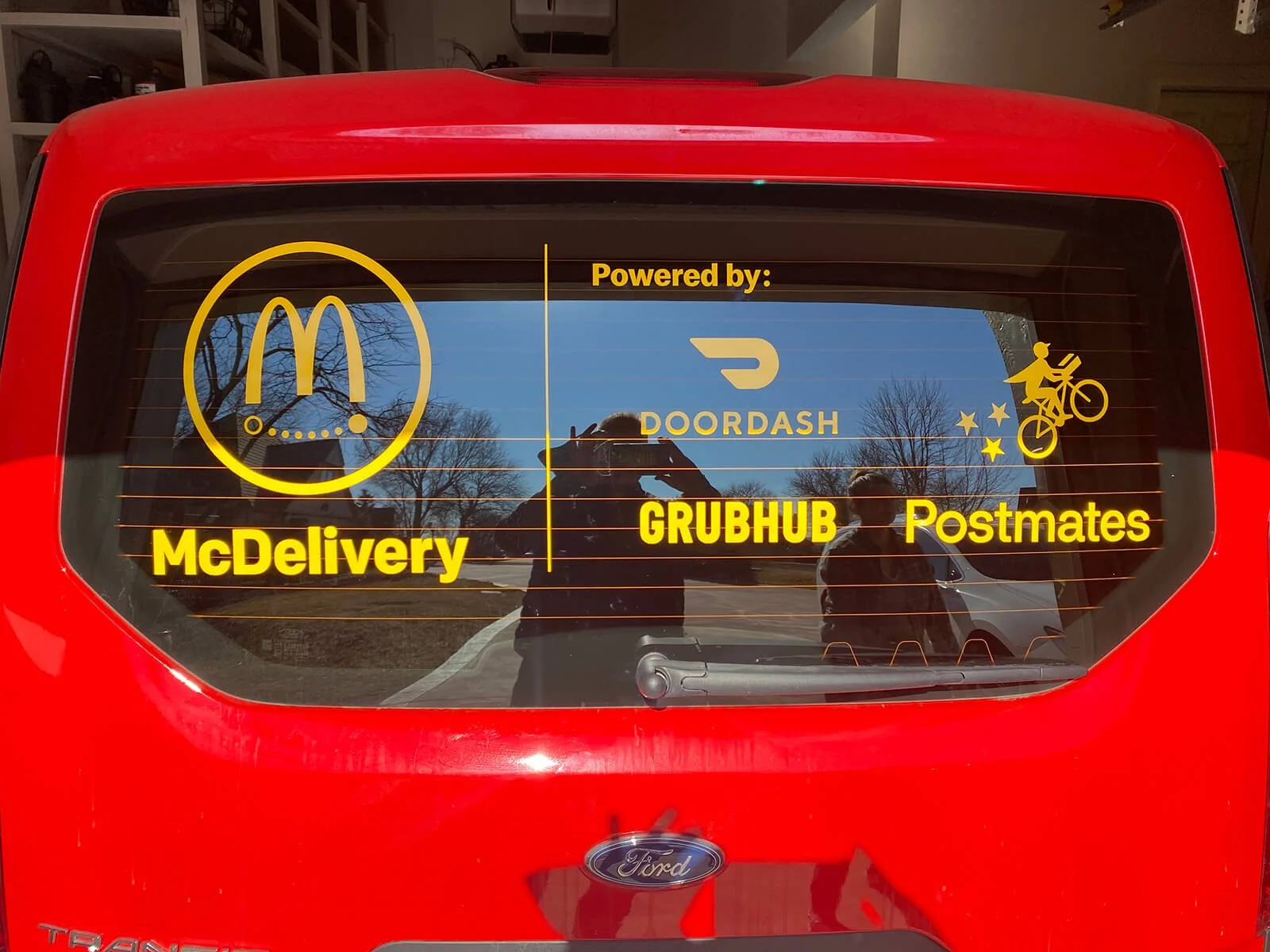 Decal on the rear window of a red vehicle showing logos and text for various food delivery services, including McDelivery, DoorDash, Grubhub, Postmates, and Uber Eats, with a yellow outline of a cyclist riding a bike.