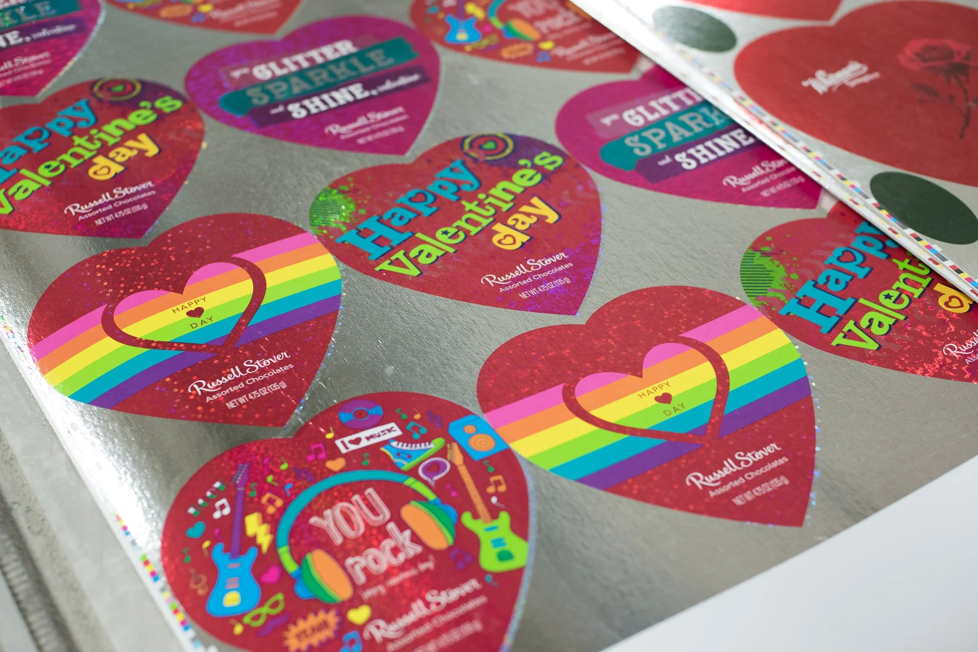 Sheet of Valentine's Day stickers with hearts featuring rainbow, musical, and glitter designs, some with the messages "Happy Valentine’s Day" and “You rock,” on a gray background.