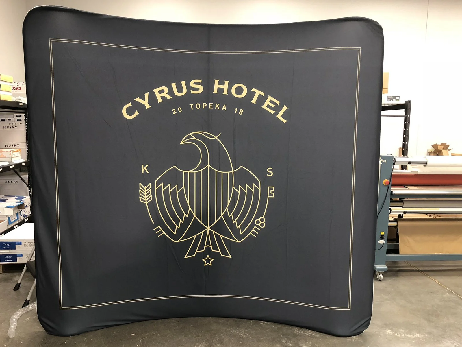Black fabric banner with gold logo and text reading "Cyrus Hotel 20 Topeka 18". The logo features a stylized eagle with outstretched wings and a shield on its chest, surrounded by the letters "K" and "S" and a small star below.